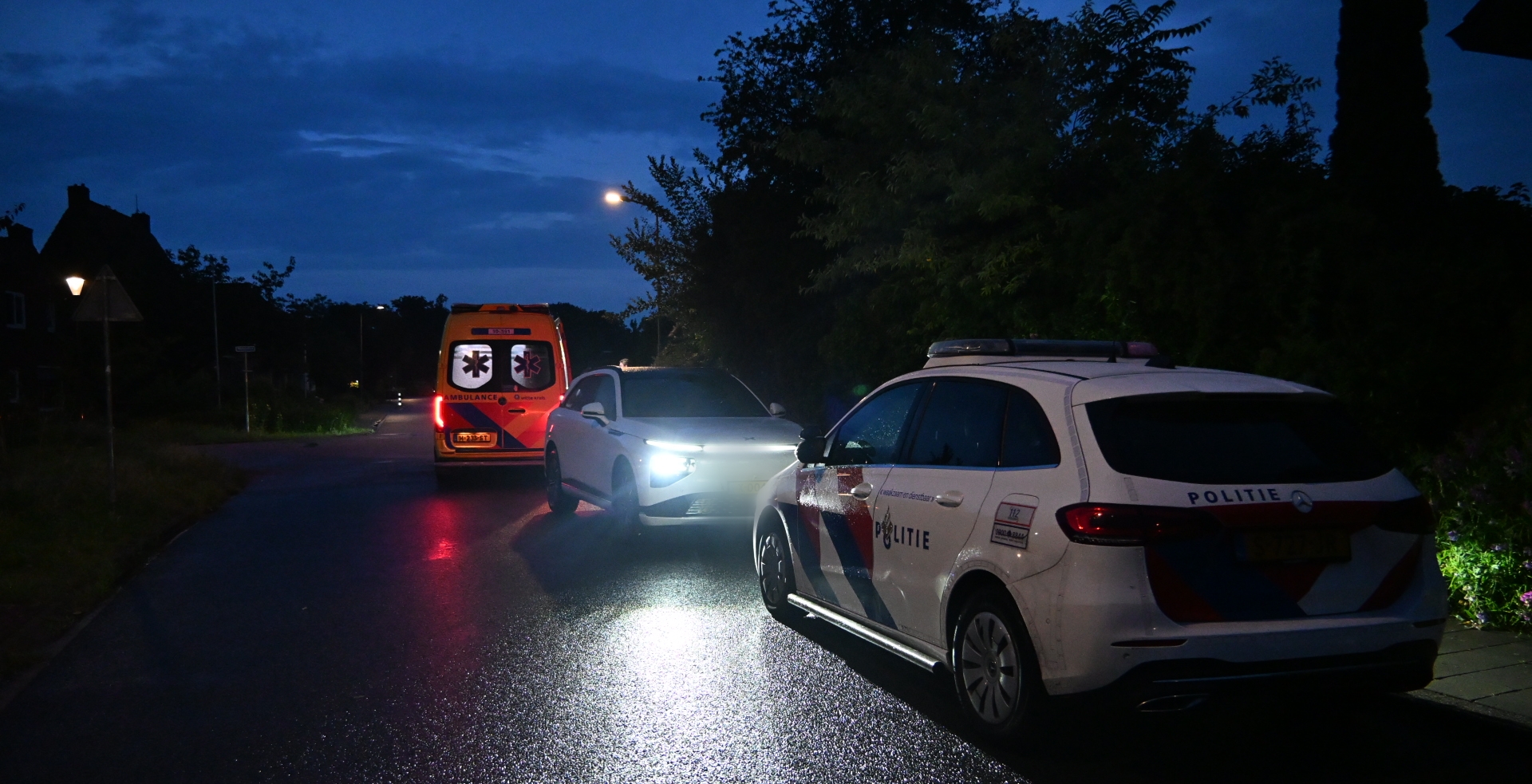 Een politieauto en een ambulance staan 's nachts op een donkere weg geparkeerd.