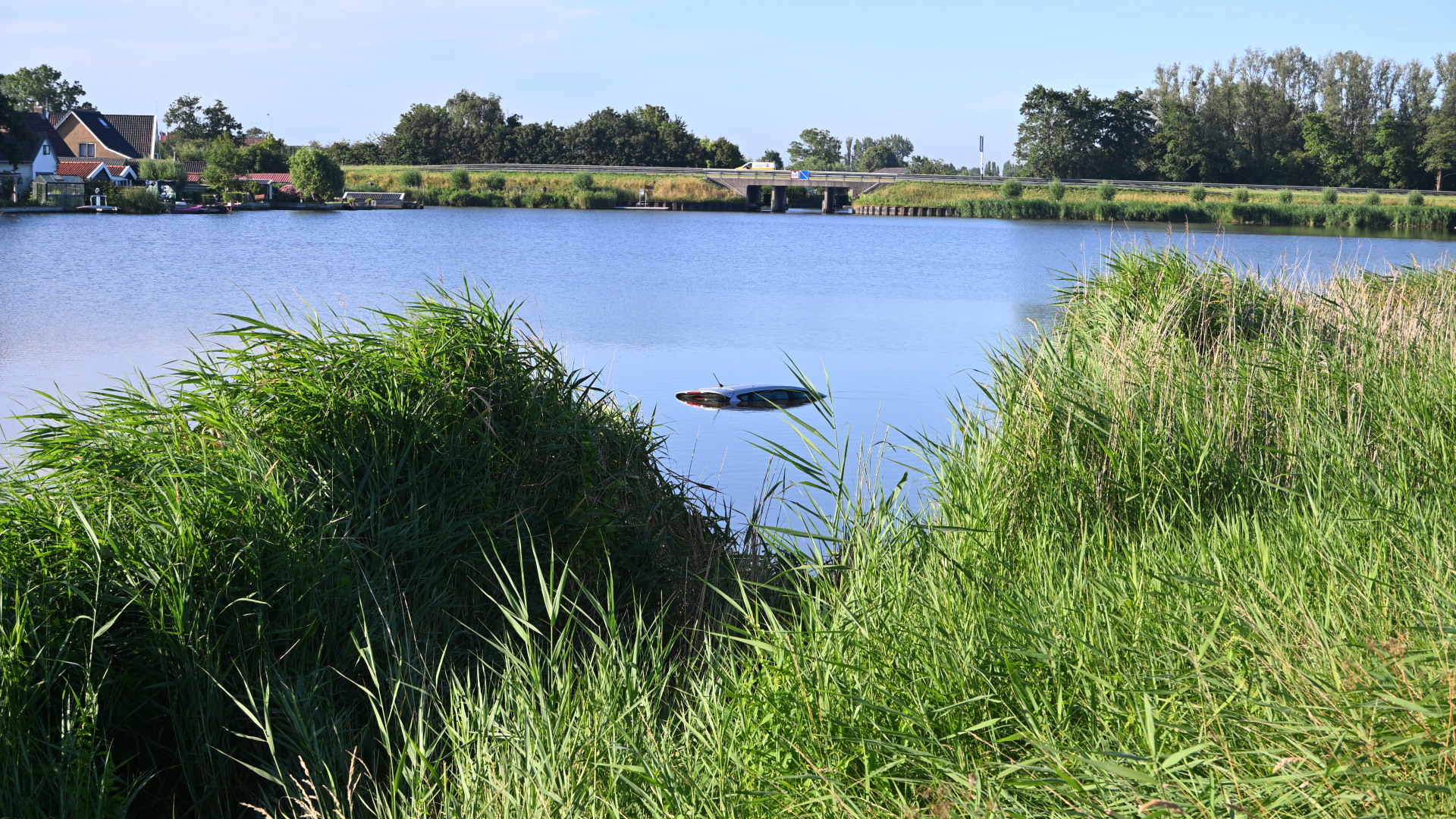 Half gezonken auto in een meer, omgeven door riet en in de verte huizen en een brug.