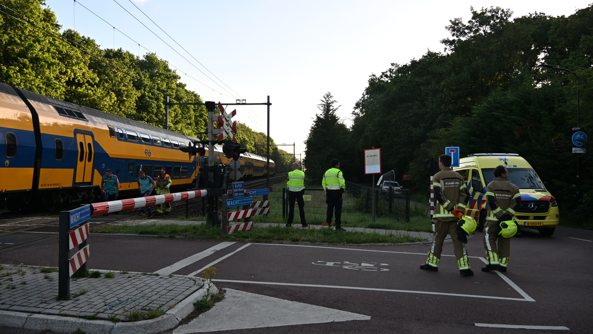 Spoorwegovergang met stilstaande trein, omgeven door politie-, ambulance- en brandweerpersoneel in gesprek.