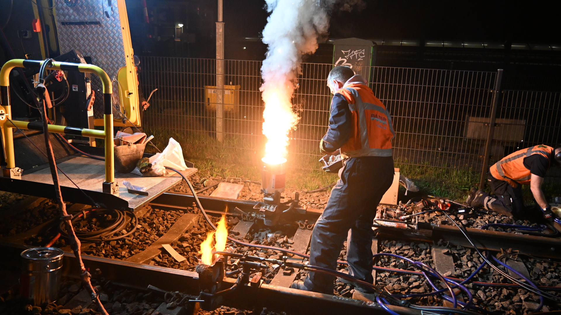 Twee spoorwerkers voeren 's nachts onderhoud uit bij een spoorlijn, waarbij vlammen uit apparatuur komen.