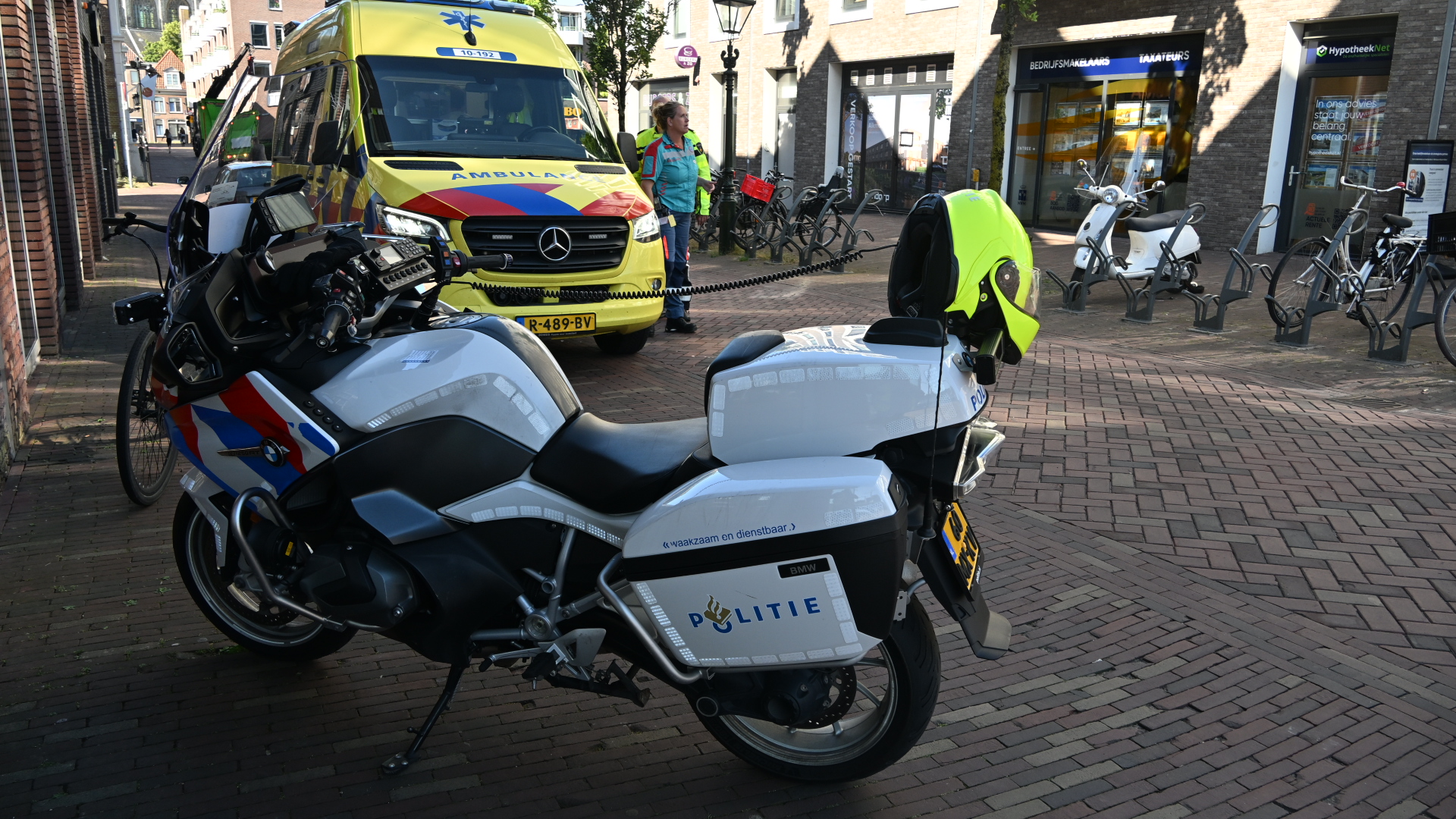 Een politie motorfiets en een ambulance staan op een plein; een medisch personeel in uniform staat dichtbij.