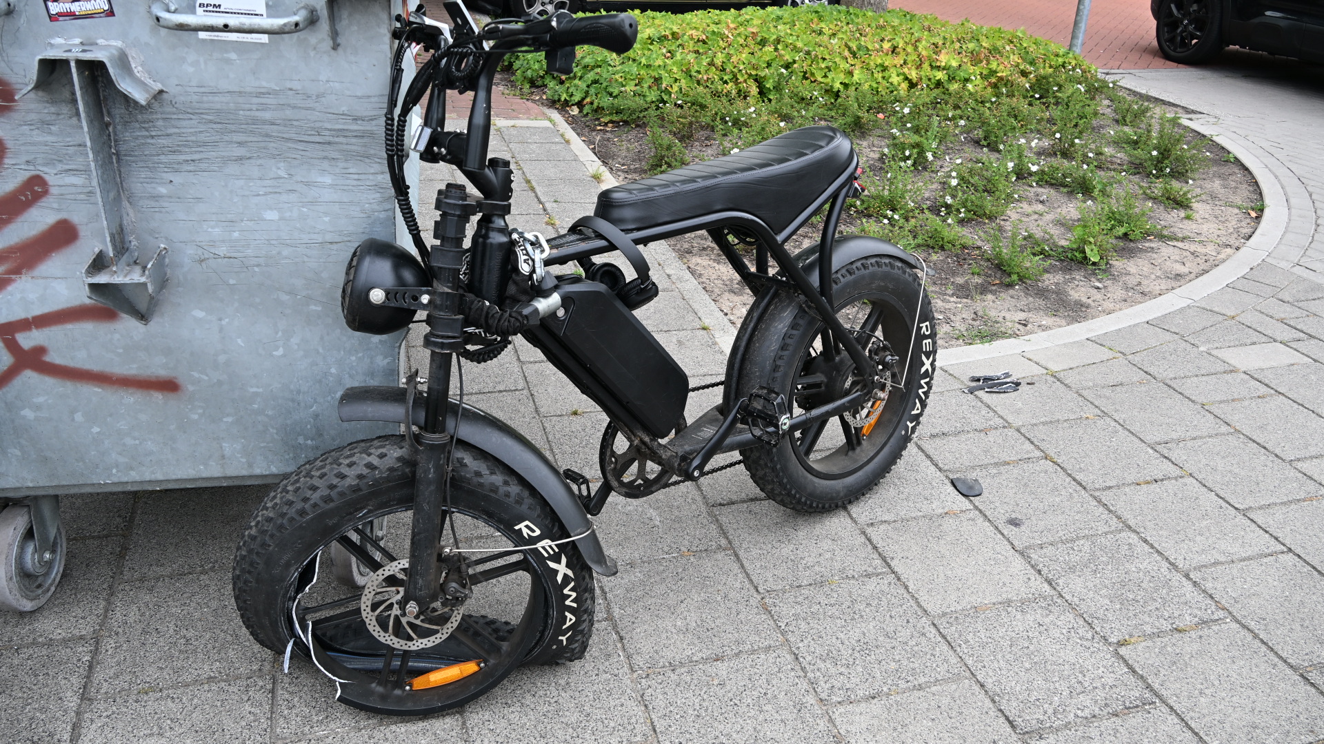 Zwarte elektrische fiets met dikke banden geparkeerd naast een metalen container op een stoep.