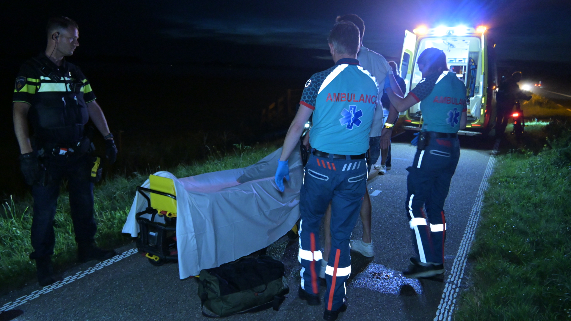 Hulpverleners en politie bij een ambulance op een donkere weg, klaar om een brancard in te laden.