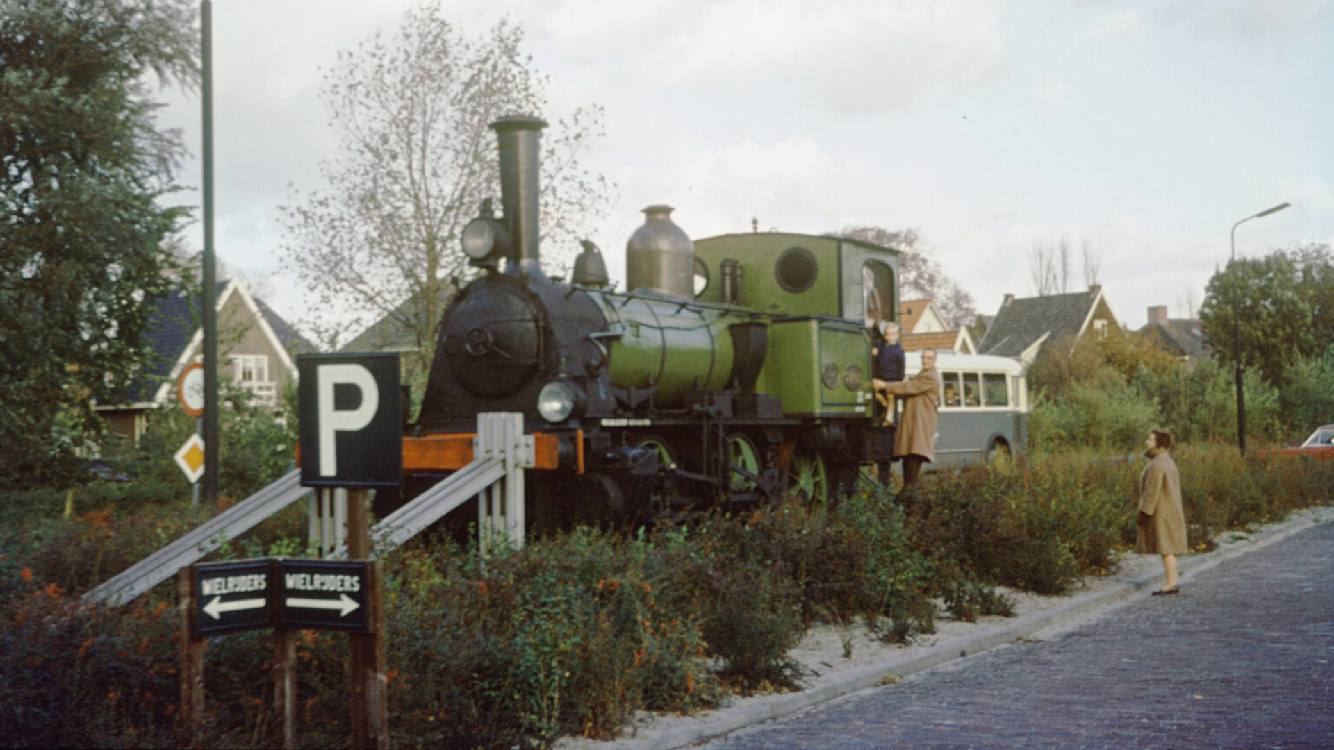 Een groene stoomlocomotief staat op een verhoogd spoor met naastgelegen borden en omringd door struiken; een ouder en kind staan erop, terwijl een vrouw ervoor kijkt.