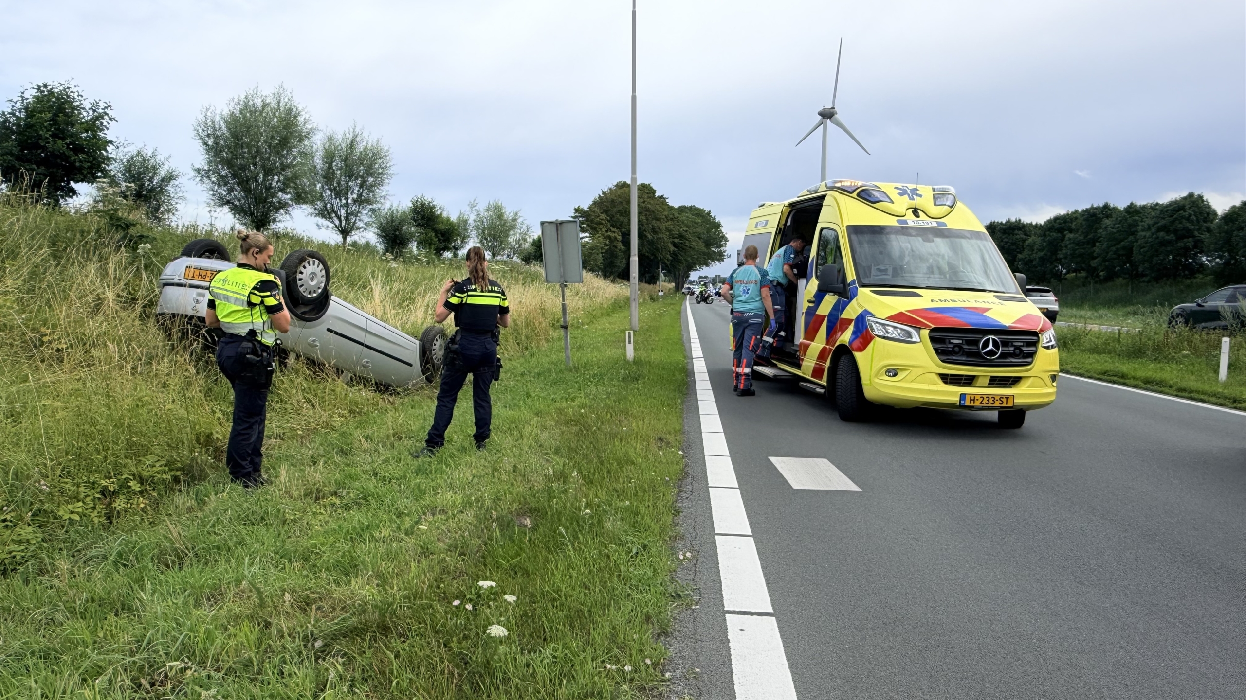 Auto ligt ondersteboven in de berm naast een weg, met politieagenten en een ambulance aanwezig.