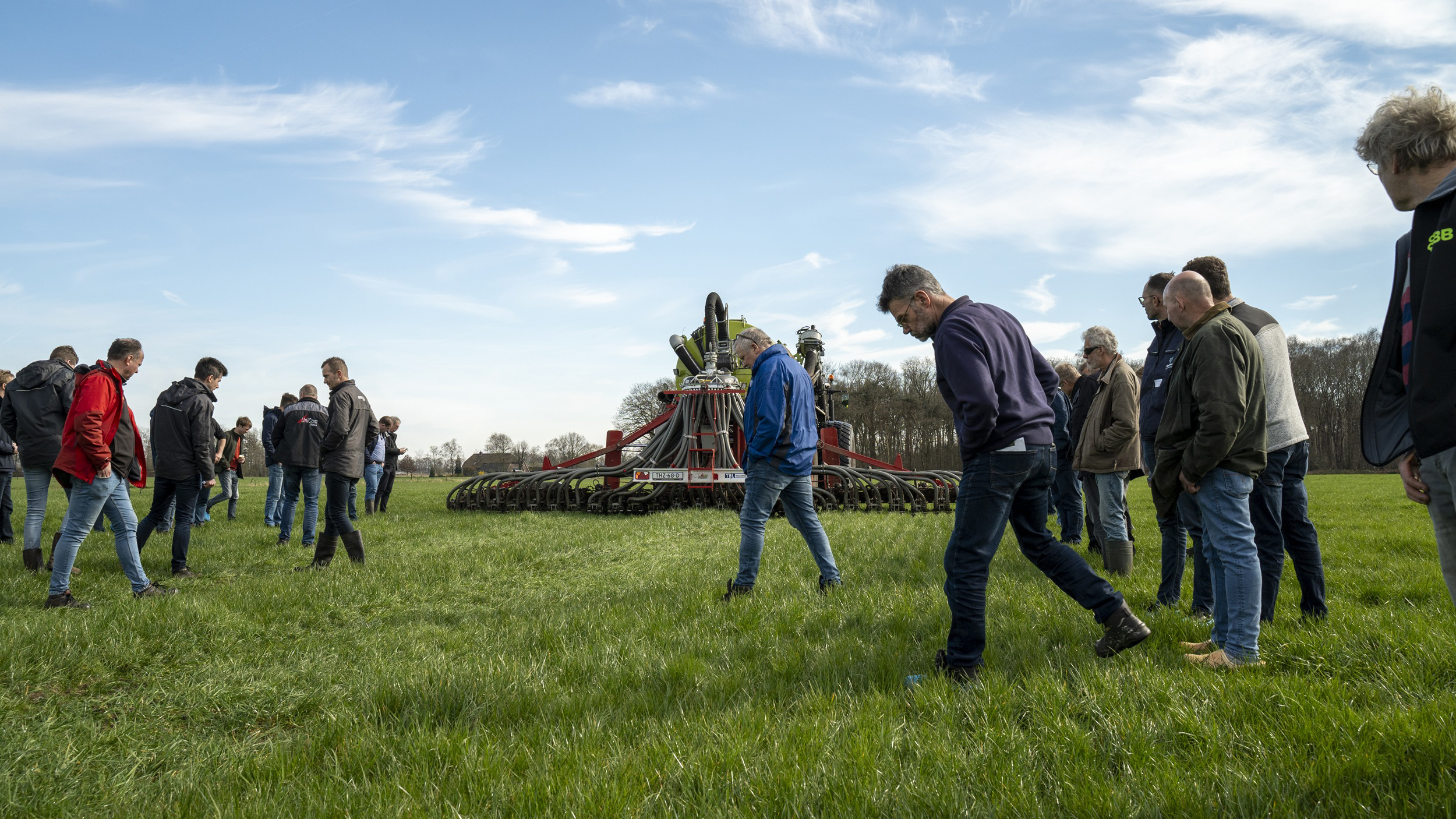 Groep mensen inspecteert een landbouwmachine op een grasveld onder een blauwe lucht.