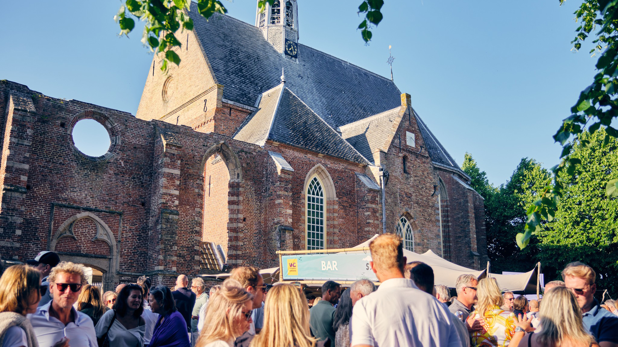 Mensen staan samen op een markt bij een oude kerk, met kraampjes en zomerse sfeer.