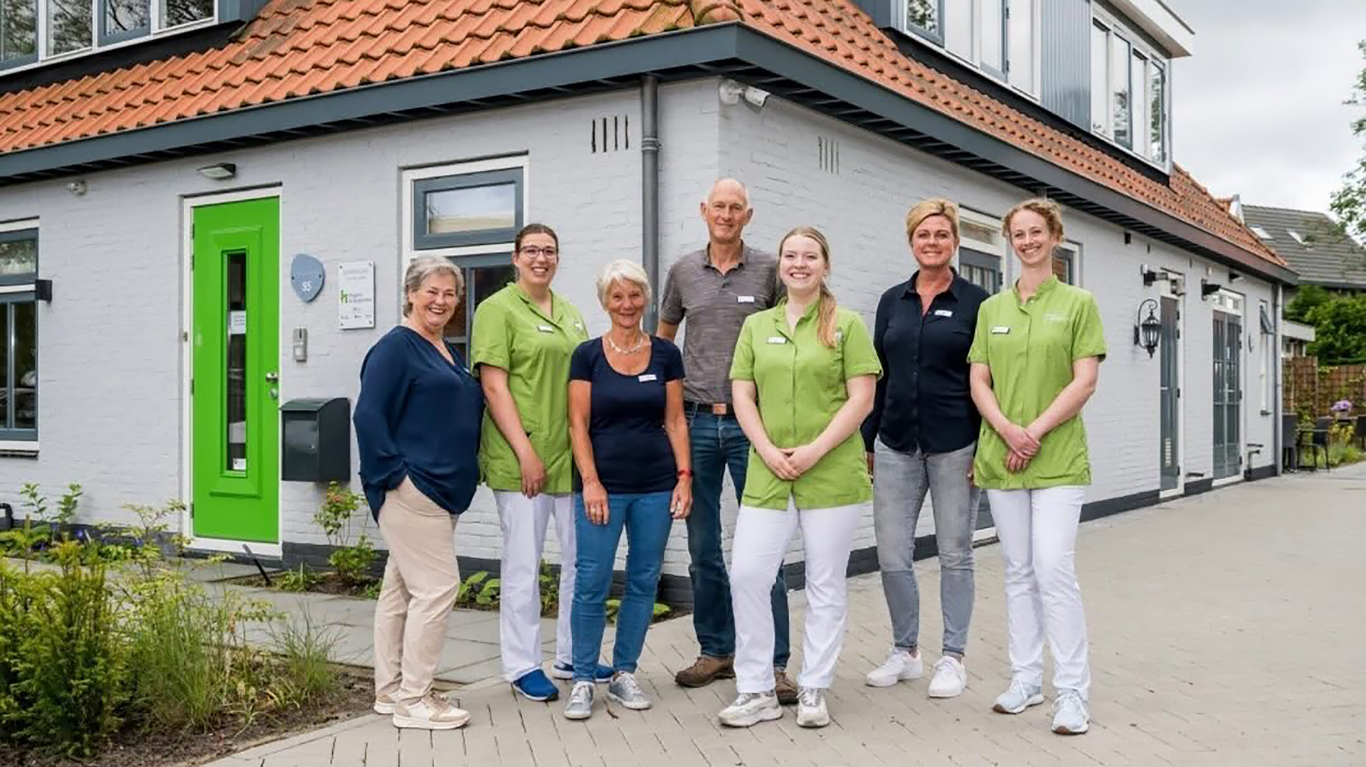 Een groep van zeven mensen poseert lachend voor een wit gebouw met een opvallende groene deur, allemaal in casual en professionele kleding.