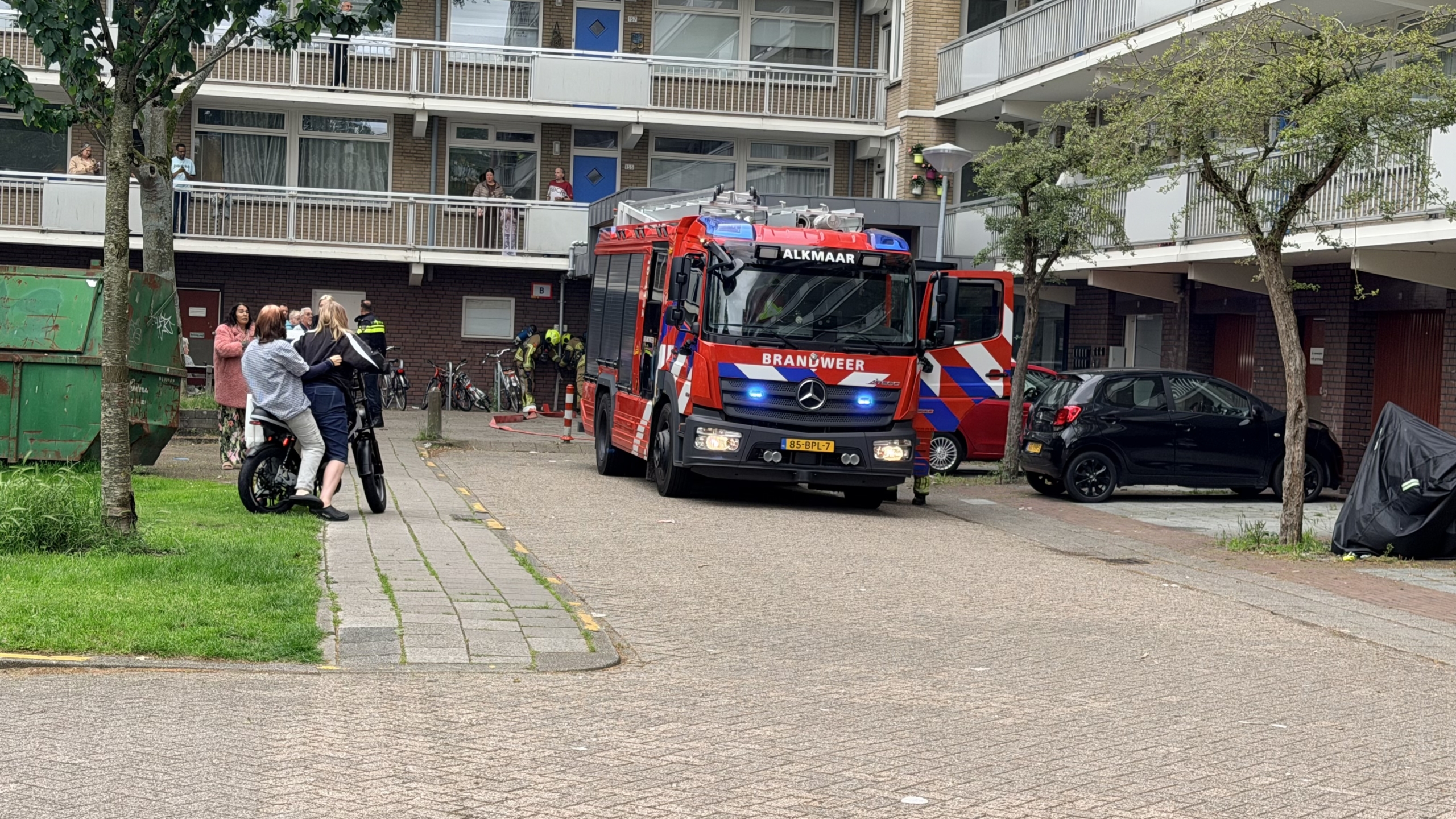Een brandweerwagen van Alkmaar staat voor een appartementengebouw, omringd door mensen en fietsen.