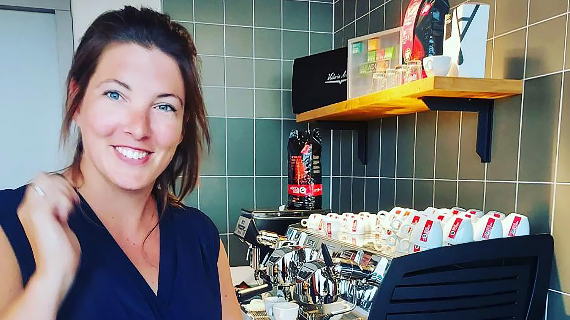 Vrouw glimlachend bij een espressomachine in een moderne keuken met glazen en koffiekopjes op een plank.