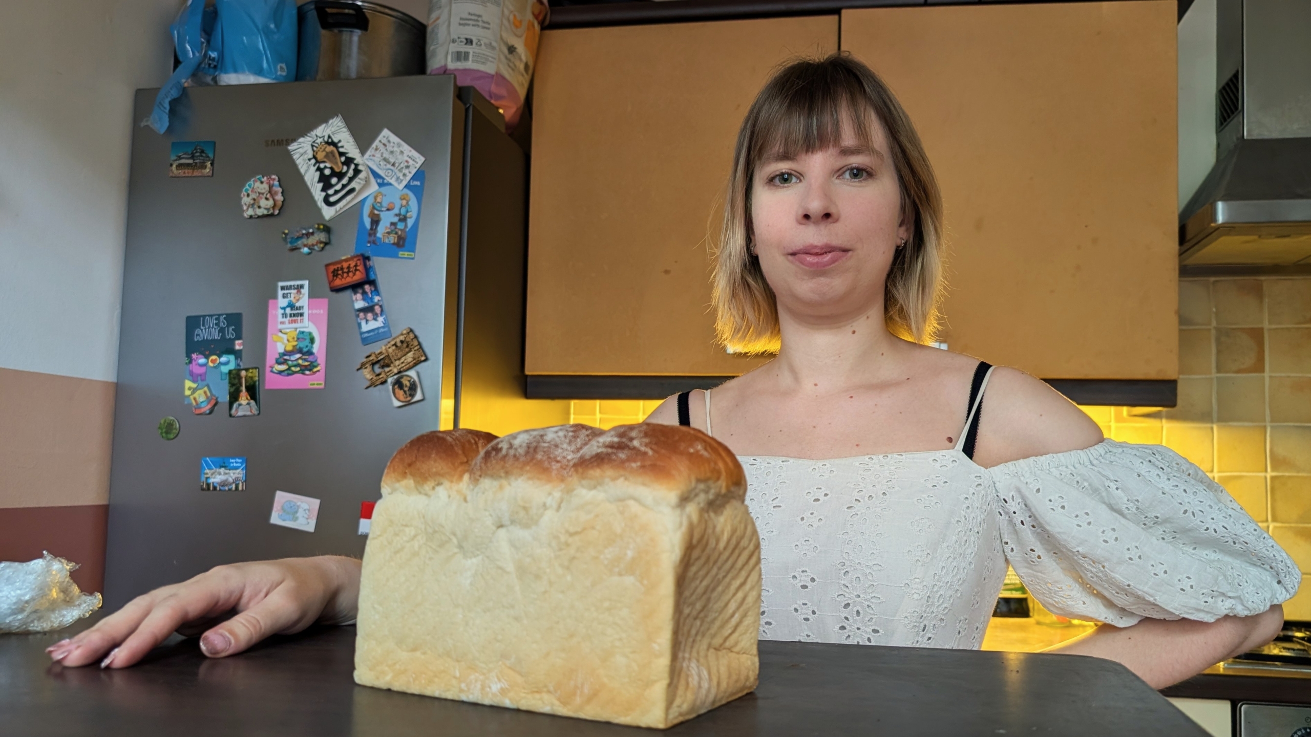 Vrouw in een keuken met een wit bloesje naast een versgebakken brood op het aanrecht, met koelkast en magneten op de achtergrond.
