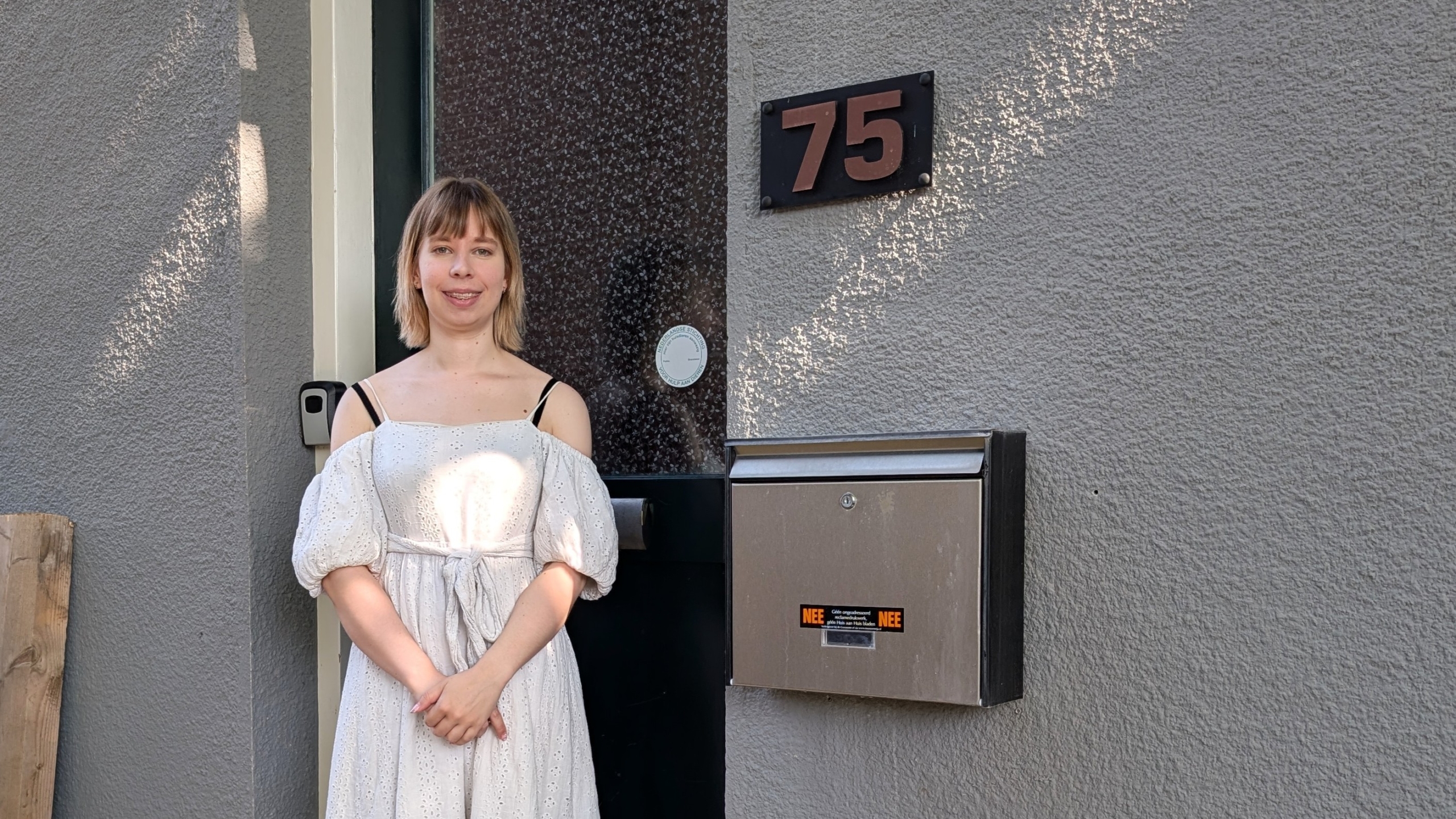 Vrouw in een witte jurk staat voor een grijze muur met een huisnummer 75 en een metalen brievenbus.