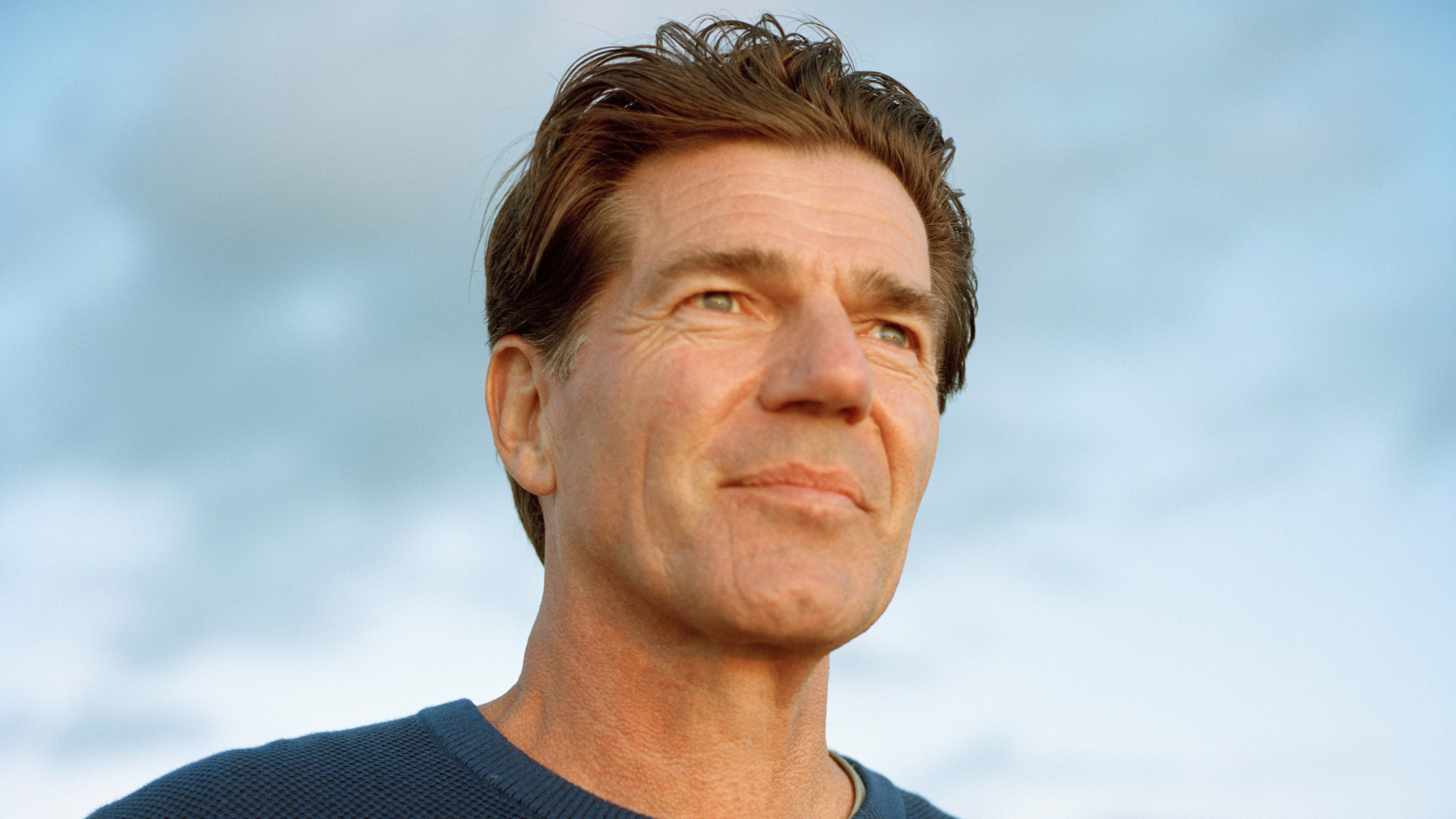 Een man kijkt in de verte tegen een heldere hemel, gekleed in een blauw trui.