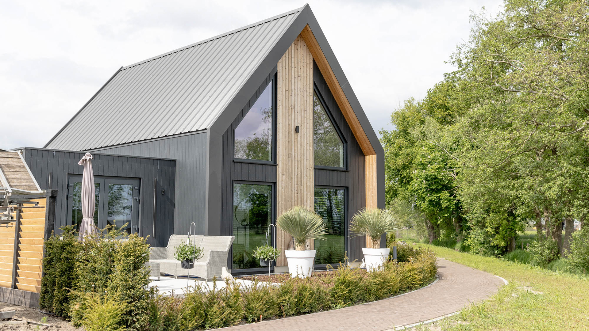 Modern huis met een zadeldak, grote ramen en houten gevelbekleding, omgeven door een tuin met planten en een terras.