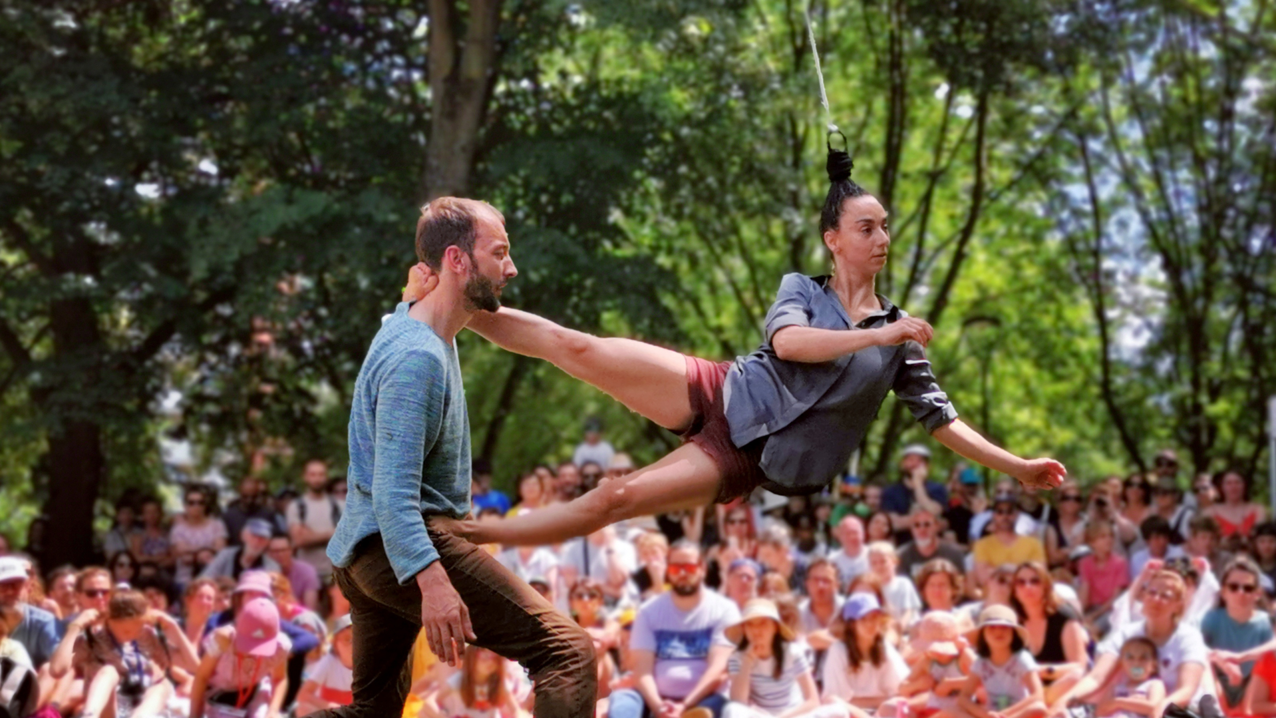Twee artiesten voeren een acrobatische act uit in een park met een publiek op de achtergrond.