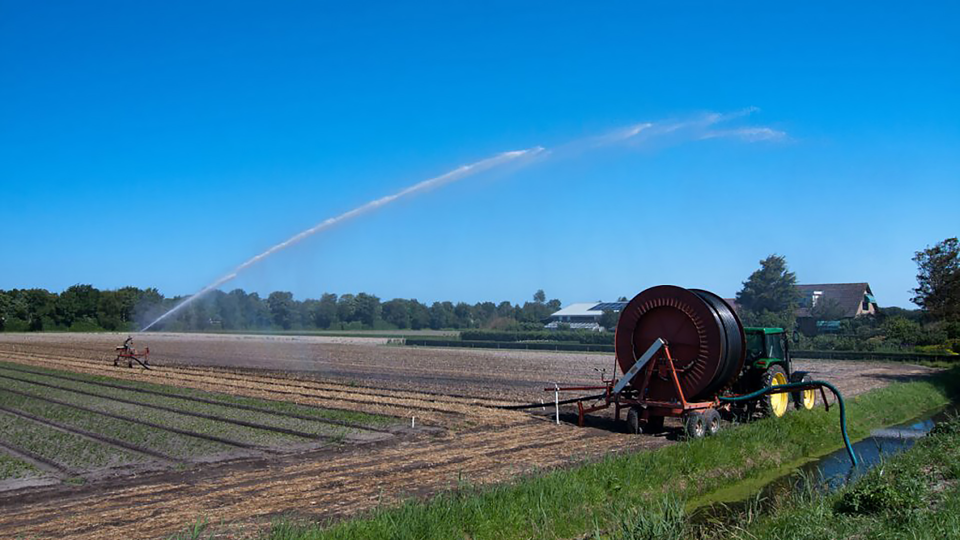 Een landbouwtractor met een sproei-installatie besproeit een groot veld onder een helderblauwe hemel.