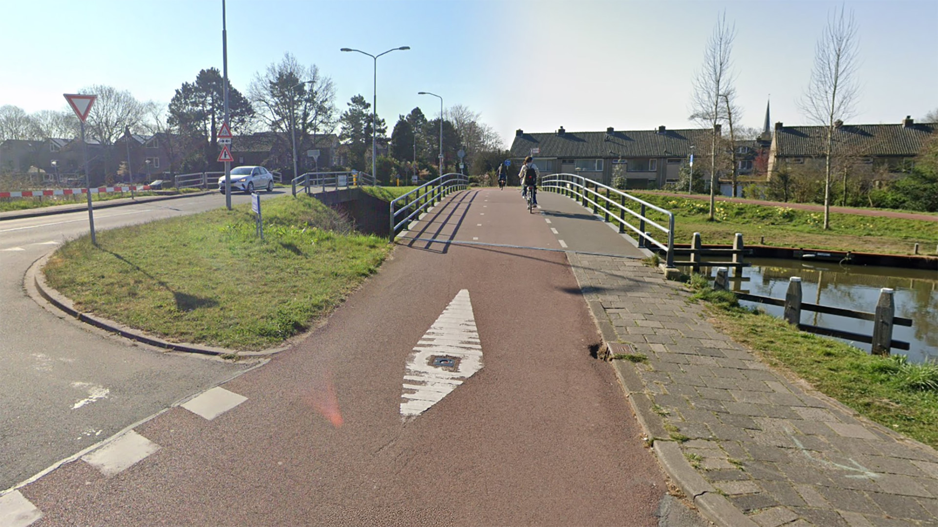 Een fietspad met een brug over een smal kanaal, twee fietsers passeren en verkeersborden staan langs de weg.