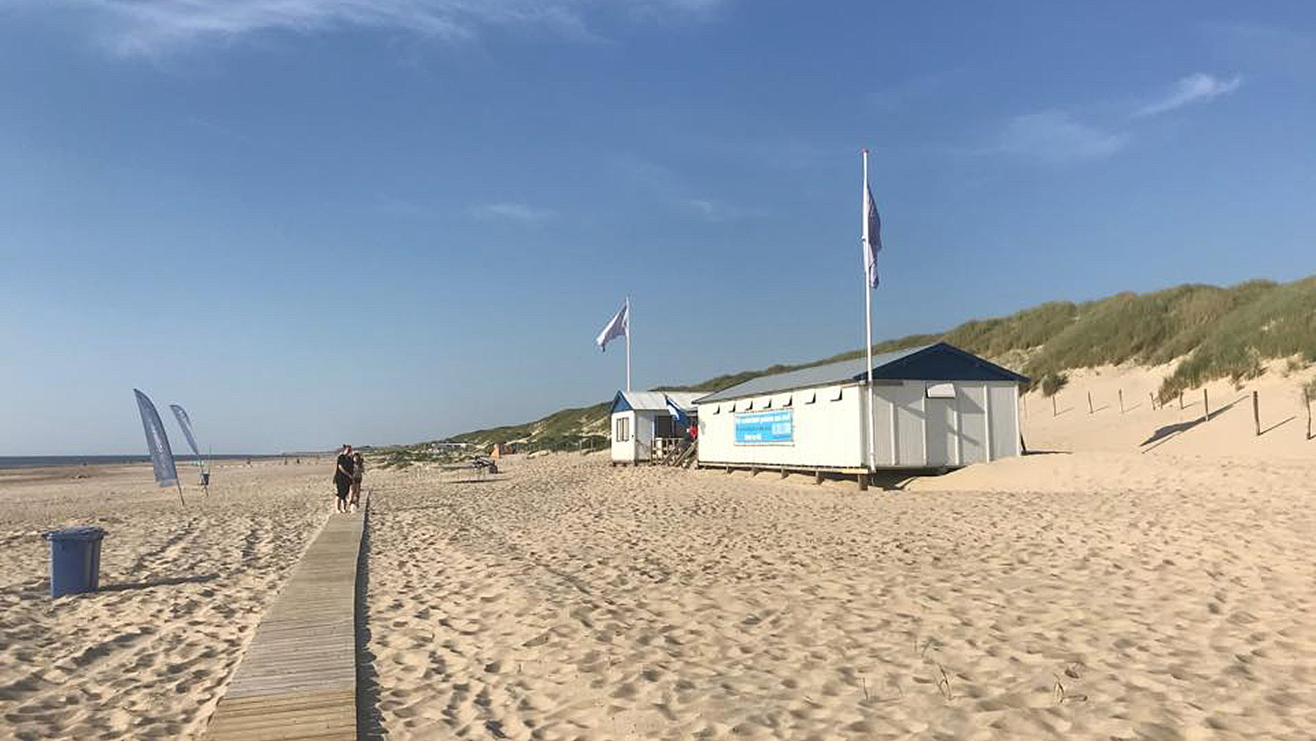 Strand met strandpaviljoen, houten pad, vlaggen, duinen en een helderblauwe lucht.