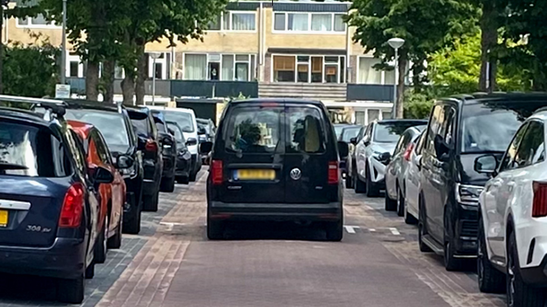 Zwarte bestelwagen geparkeerd in een smalle straat met auto's aan weerszijden en appartementen op de achtergrond.