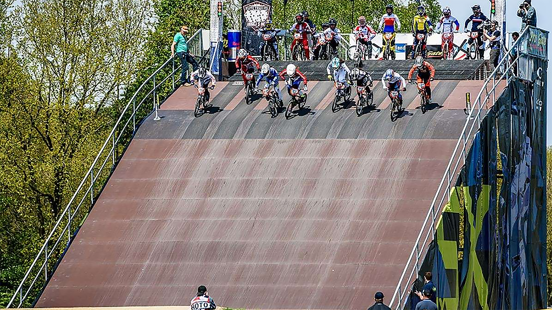 BMX-rijders starten vanaf een hoge helling tijdens een wedstrijd, omringd door toeschouwers en officials.