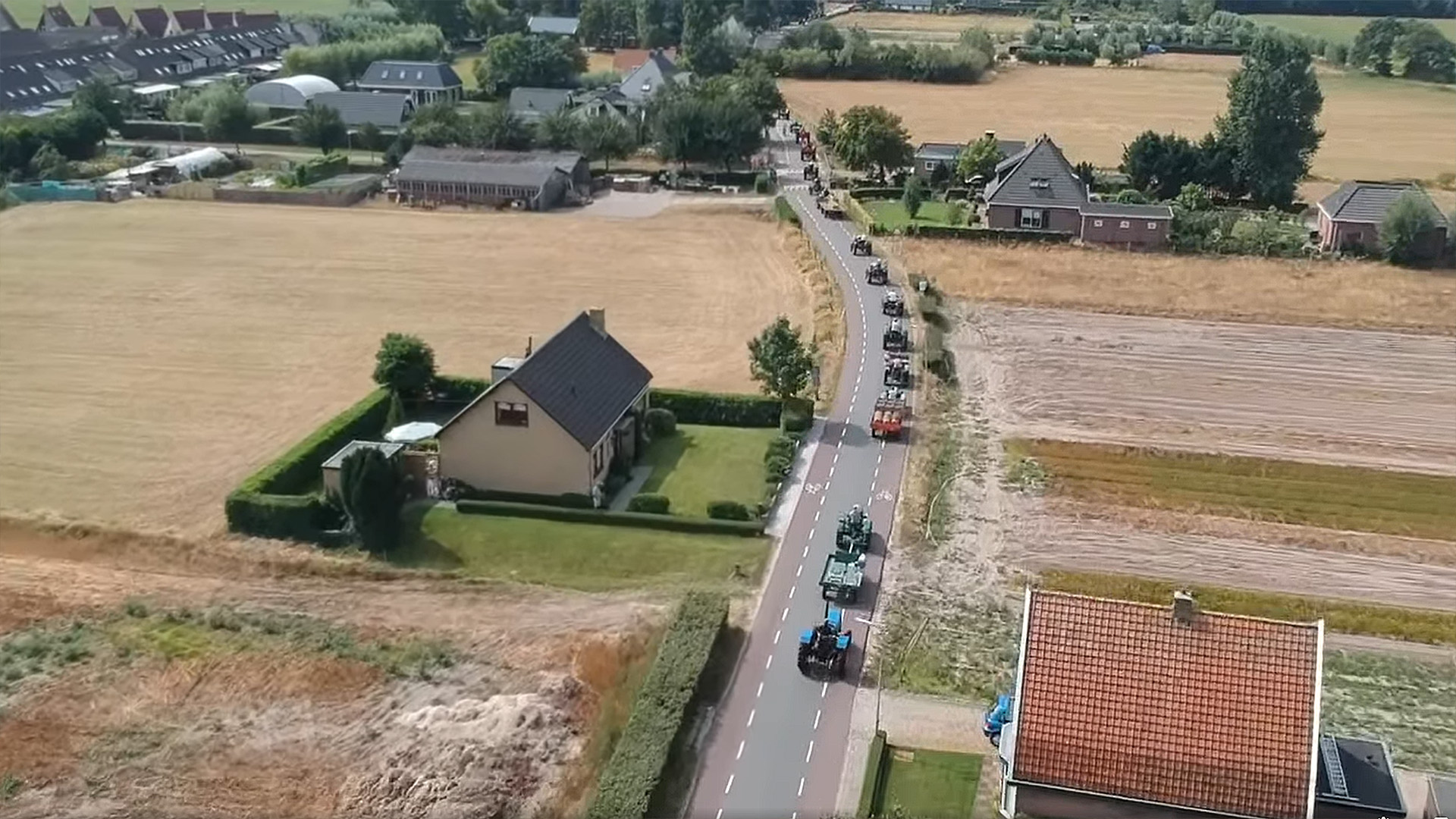 Luchtfoto van een tractorstoet op een landweg langs akkers en boerderijen in een dorp