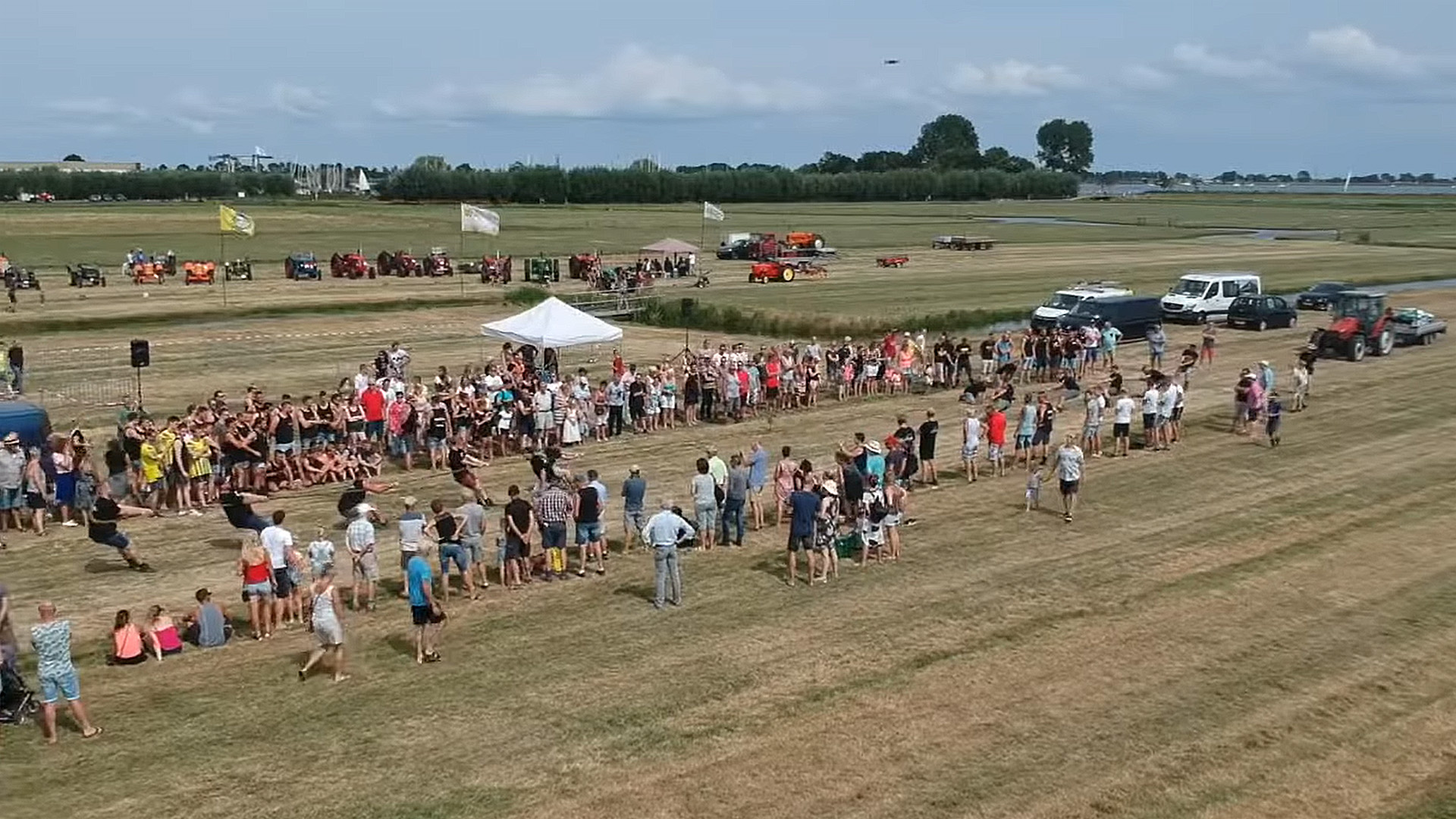 Grote groep toeschouwers op een grasveld bij een touwtrekwedstrijd, met tractoren en tentjes op de achtergrond