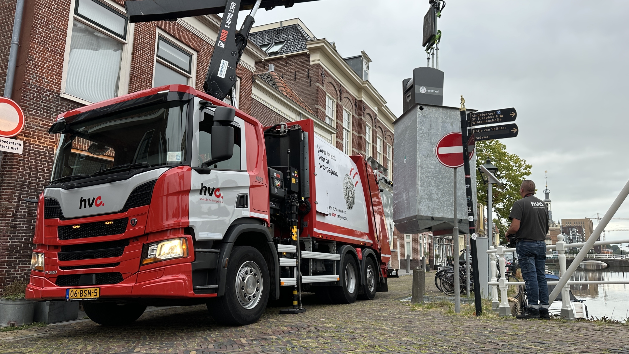HVC-vuilniswagen met kraan tilt ondergrondse afvalcontainer naast kanaal, met medewerker en verkeersborden op de kade