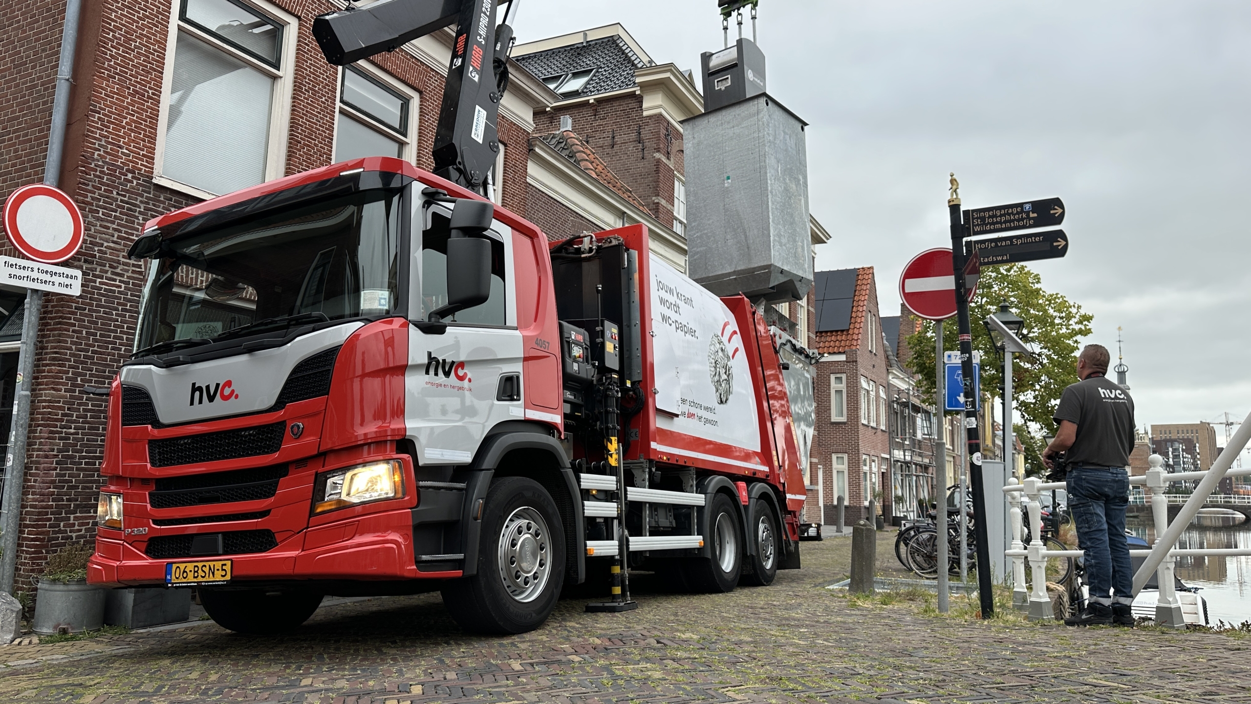 Rode HVC-vrachtwagen met kraan hijst grijze container in smalle straat langs gracht, medewerker staat ernaast