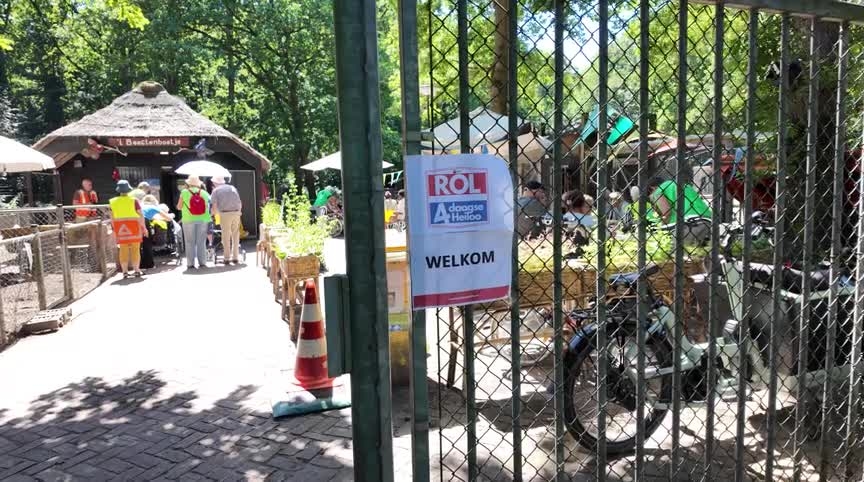 Een hek met een welkomstbord opent naar een park met mensen bij een ingang, omringd door bomen en tafels.