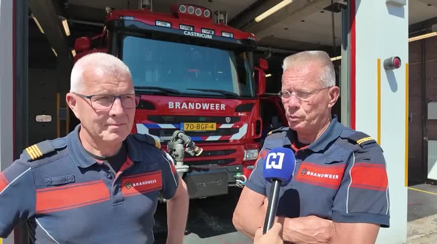 Twee brandweermannen worden geïnterviewd voor een brandweerwagen met opschrift Brandweer bij een kazerne