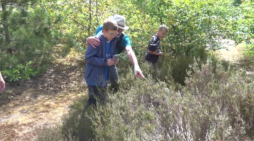 Twee mensen in de struiken in een bos; een man wijst iets aan terwijl de ander op een telefoon kijkt, met een kind erachter
