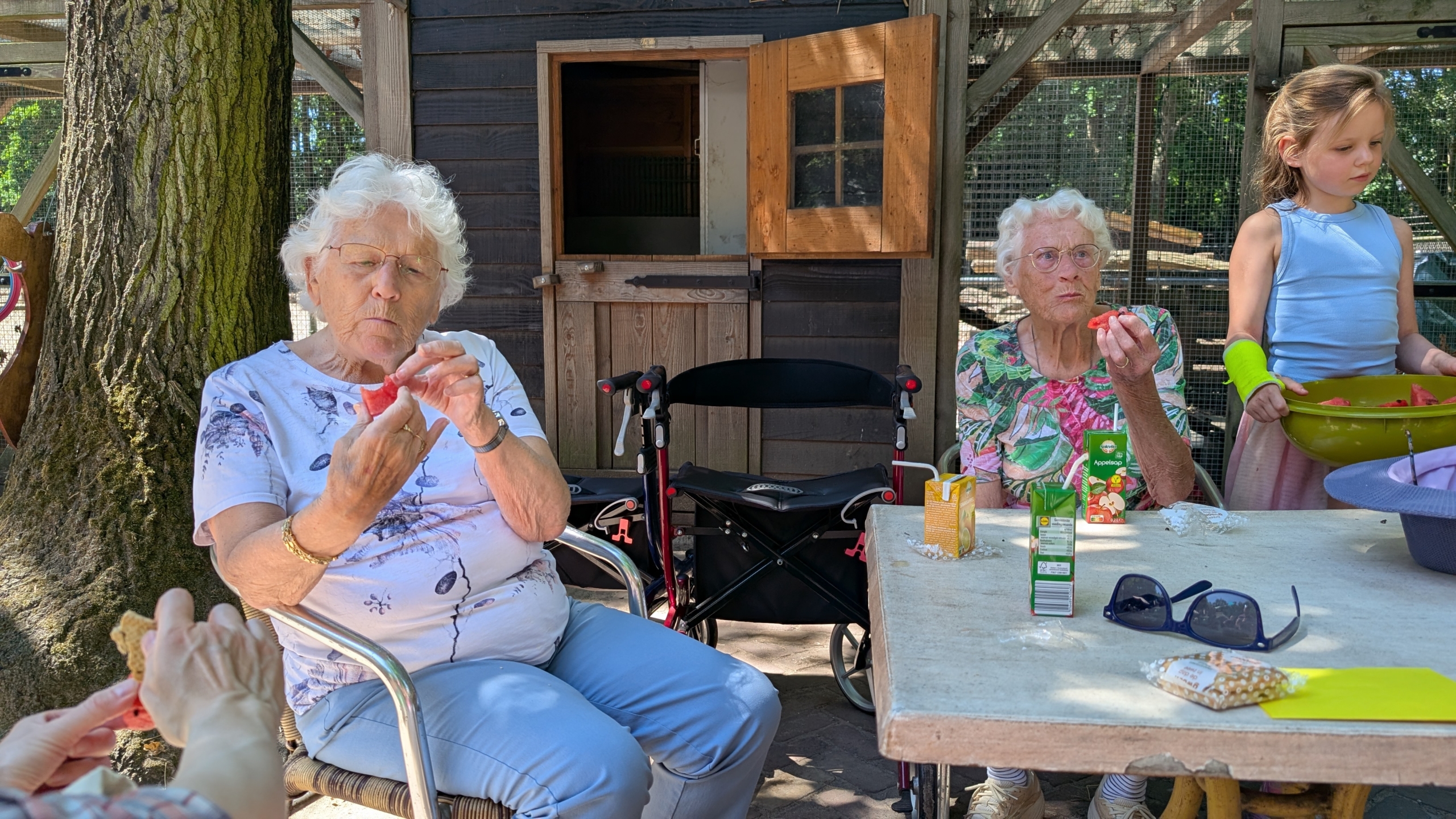 Oudere vrouwen en een meisje eten watermeloen aan een tafel buiten, bij een houten schuur.