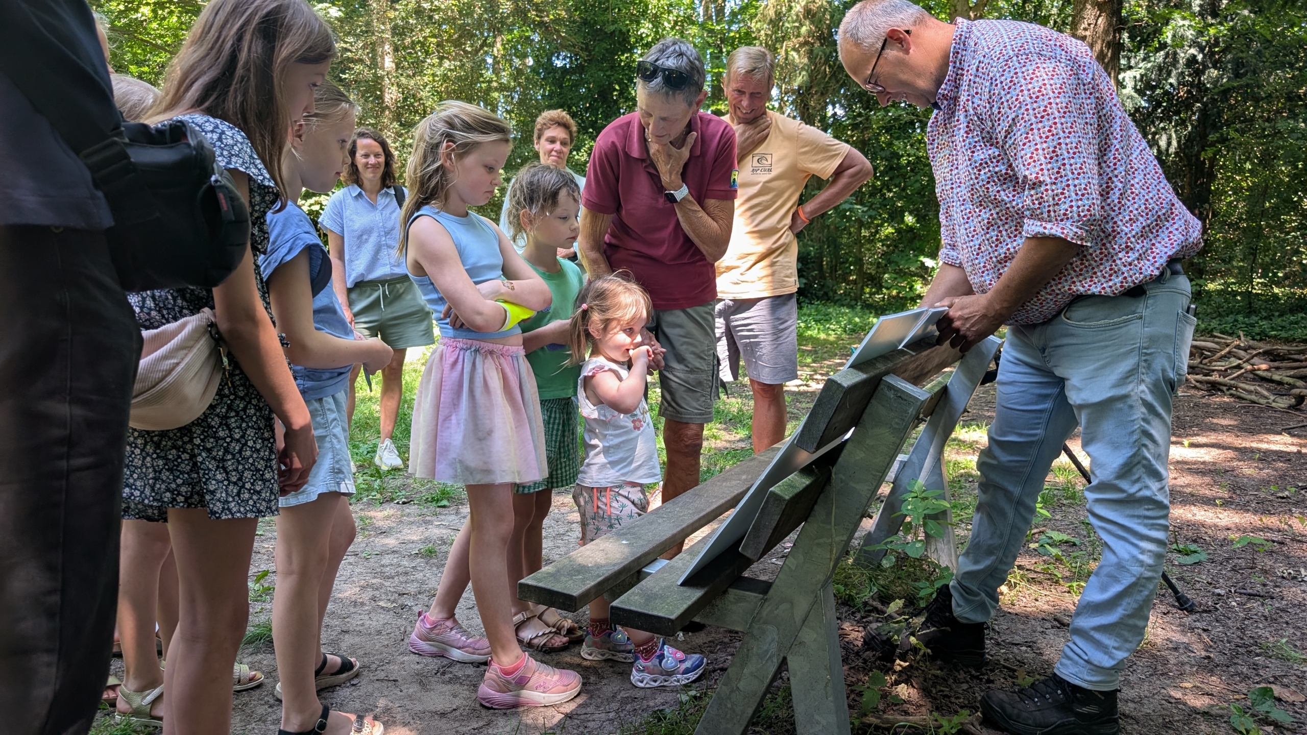 Een groep kinderen en volwassenen kijkt naar een man die iets op een bank in het bos laat zien.