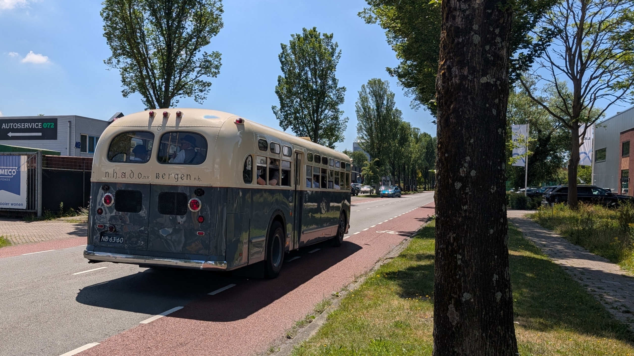Klassieke bus met opschrift "n.h.a.d.o nv bergen" rijdt over een weg omgeven door bomen en gebouwen.