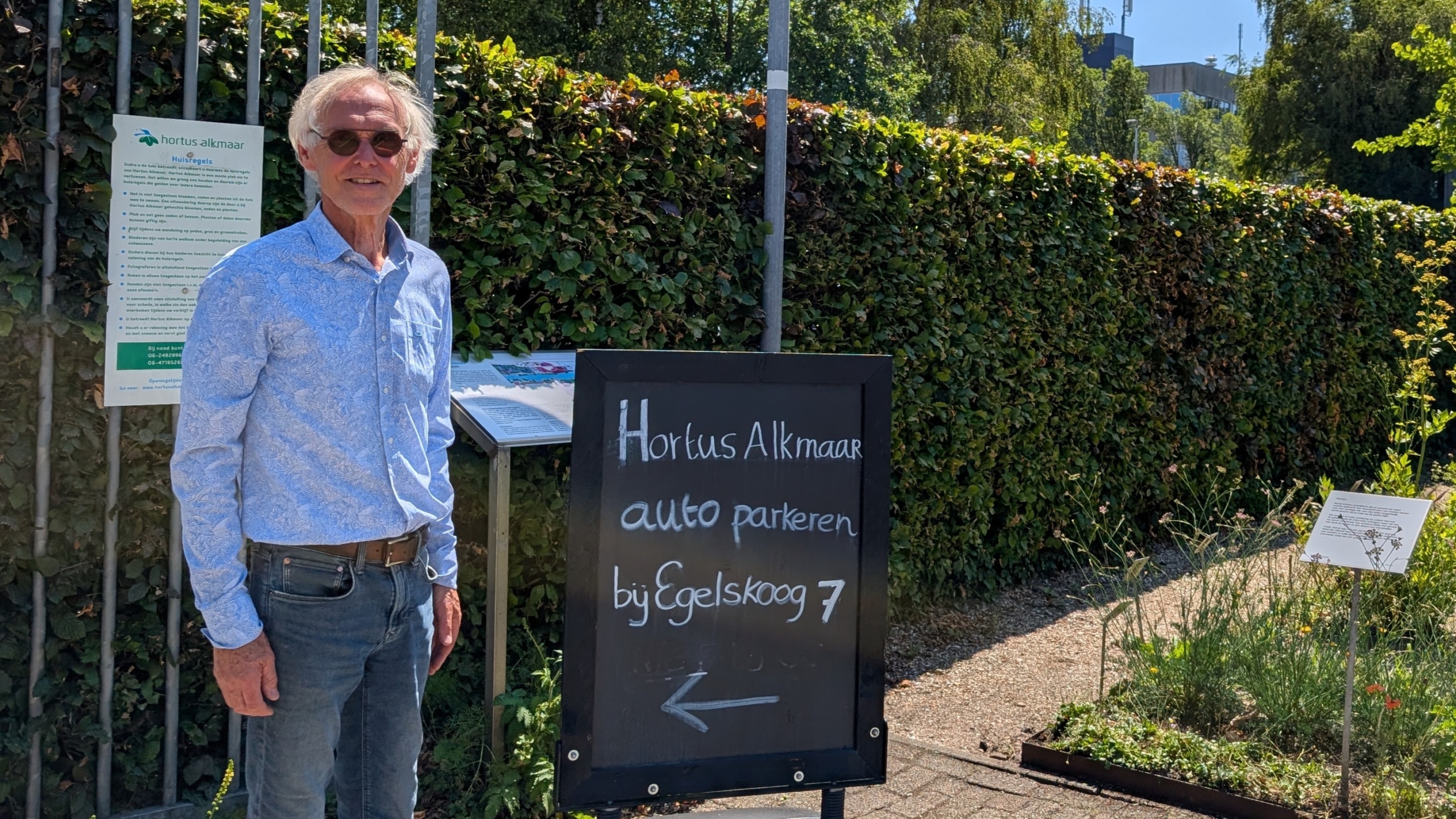 Man in een blauwe blouse naast een bord met de tekst "Hortus Alkmaar auto parkeren bij Egelskoog 7" in een groene tuinomgeving.
