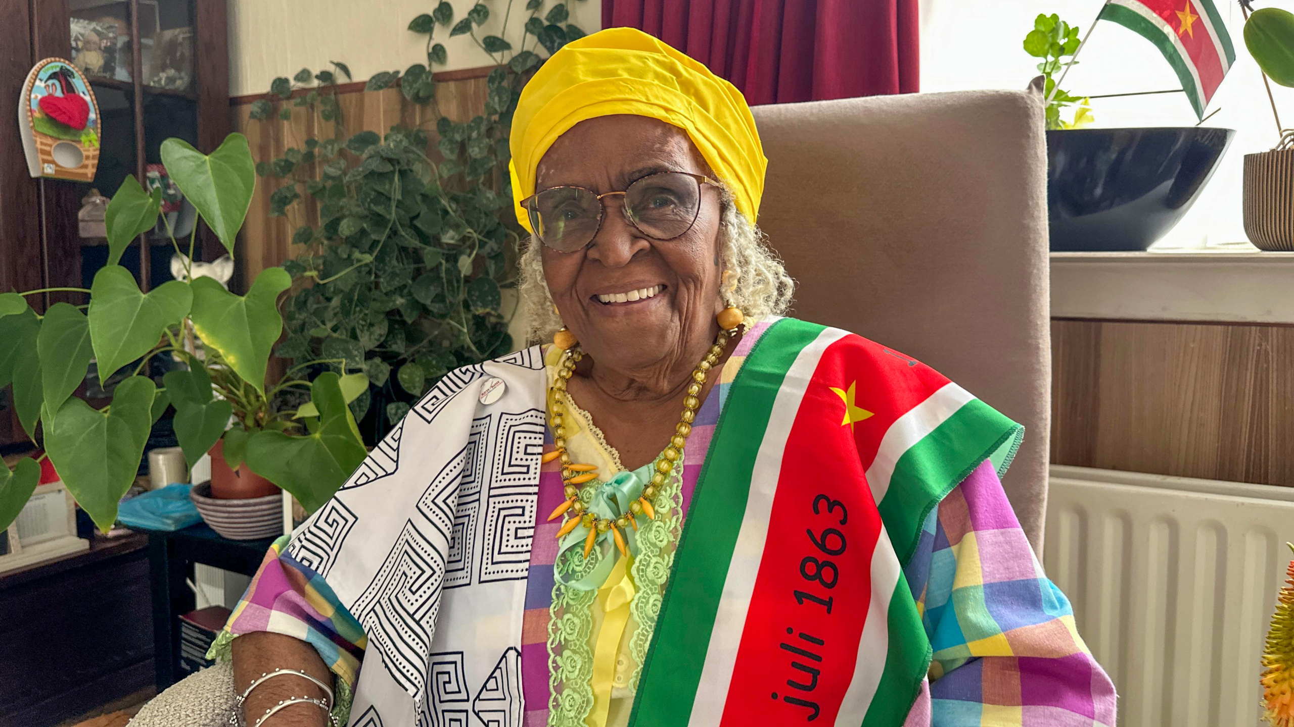 Een oudere vrouw met een gele hoofddoek en kleurrijke kleding zit glimlachend naast een plant, met een Surinaamse vlag om haar schouder.
