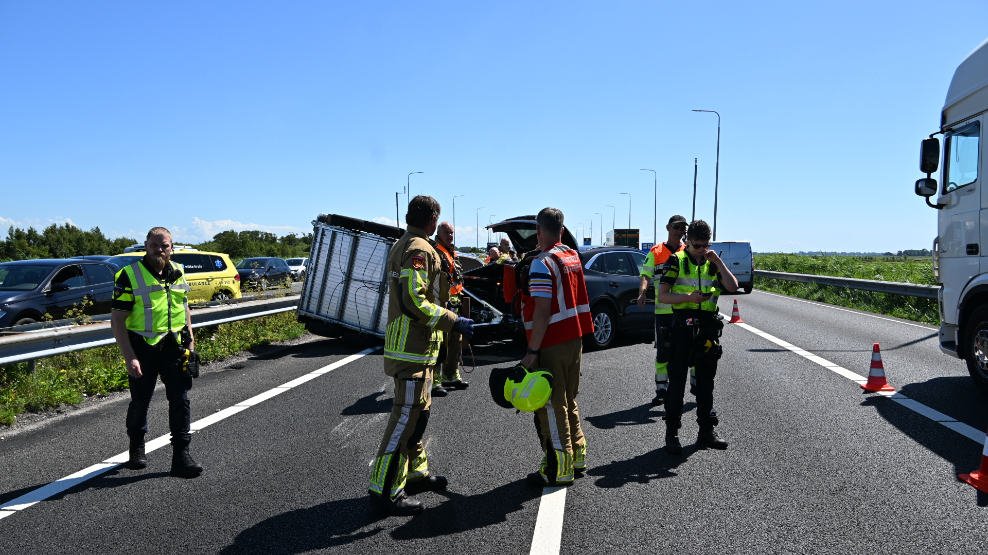 Hulpverleners op de snelweg bij een ongeval, naast een beschadigde auto en een afgebroken aanhanger.