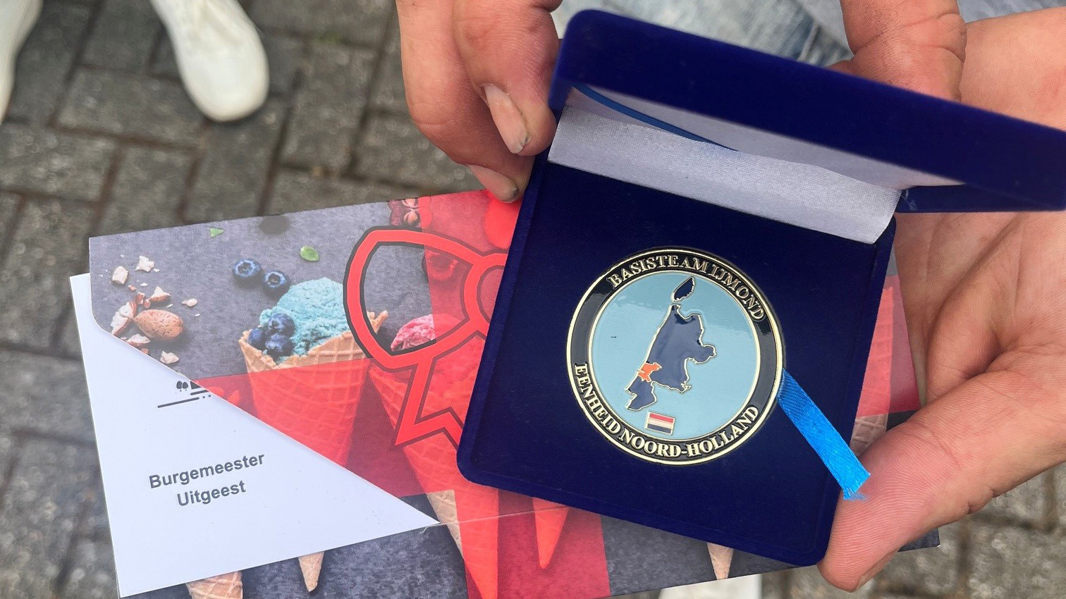 Iemand houdt een medaille in een blauw doosje met de tekst "Basisteam IJmond Eenheid Noord-Holland", naast een envelop van de burgemeester van Uitgeest.