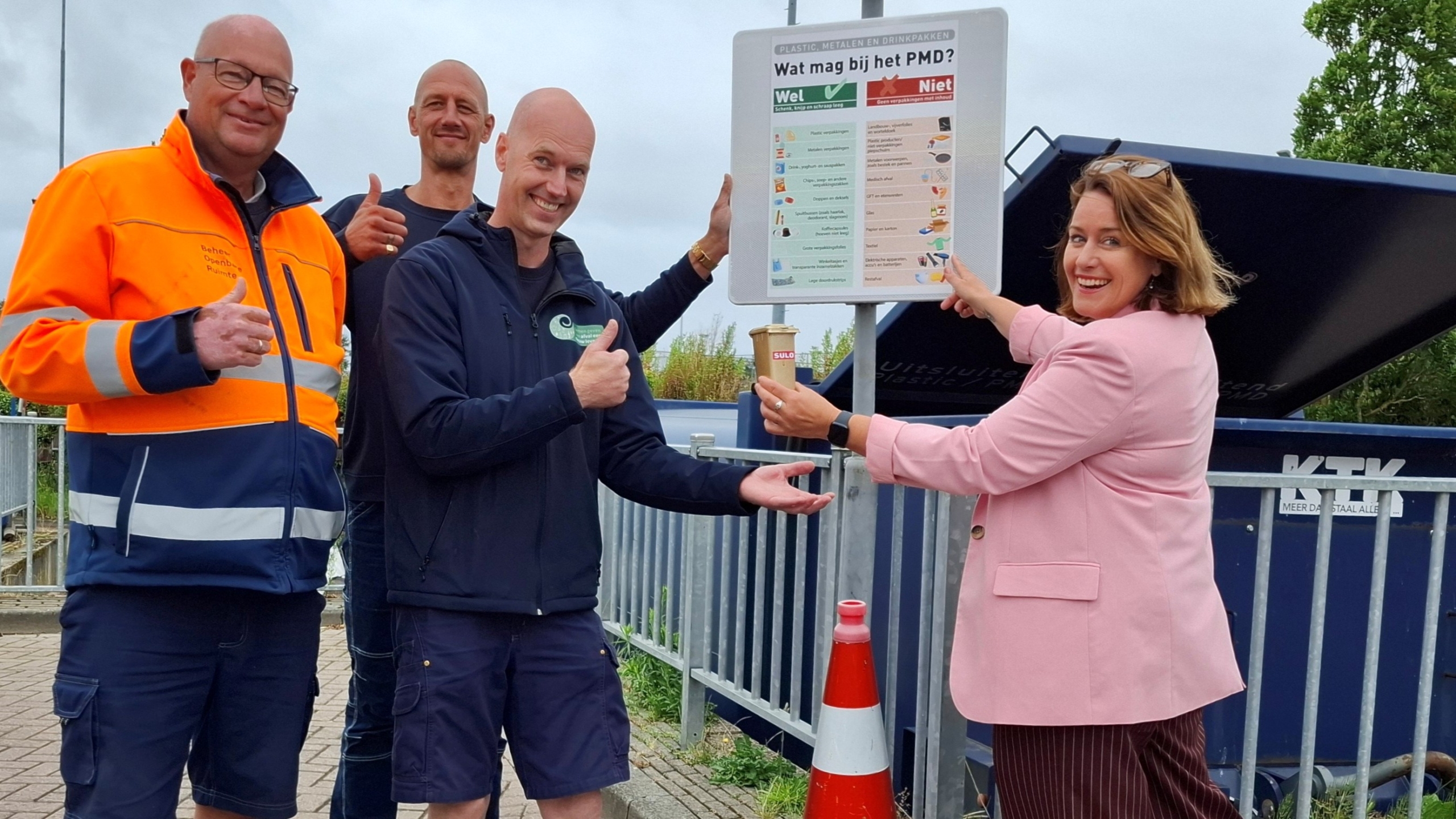 Vier mensen poseren voor een PMD-informatiebord bij een afvalcontainer, waarbij ze goedkeurend gebaren en lachen.