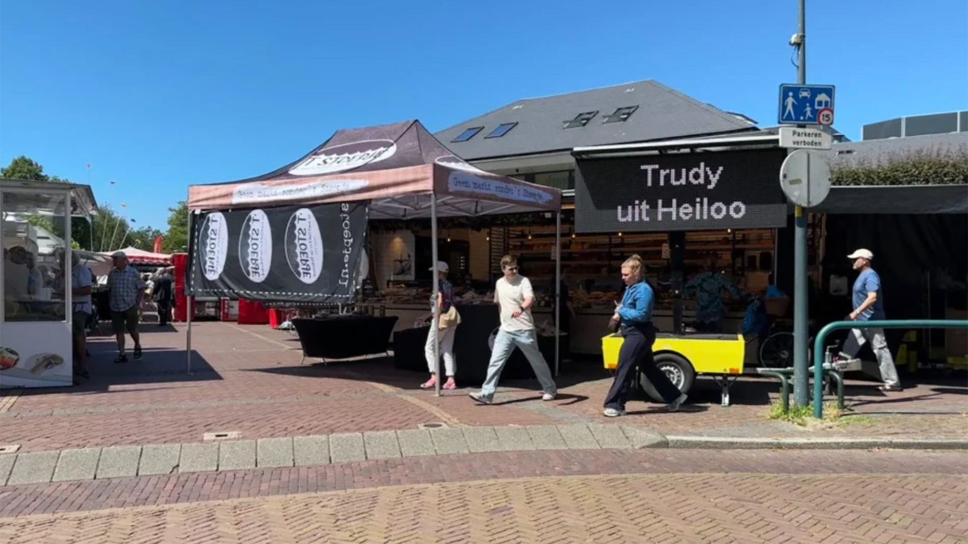 Straatscène met een markt, een kraam met "Trudy uit Heiloo" op een scherm, en mensen die voorbijlopen.