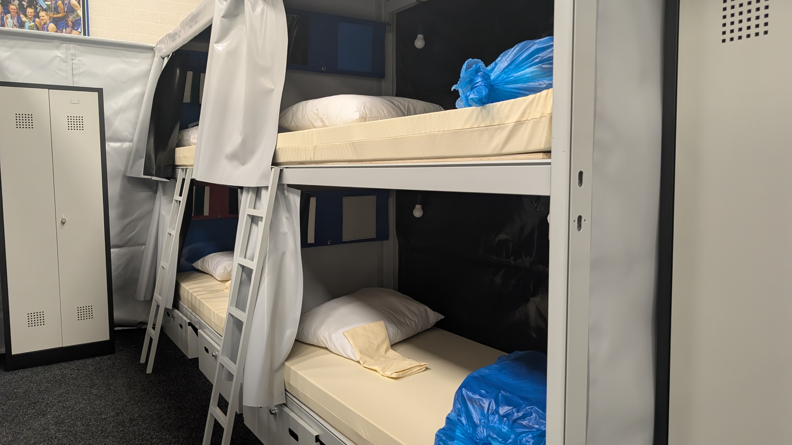 Twee stapelbedden met kussens en blauwe zakken, omgeven door gordijnen en een metalen kast in een slaapruimte.