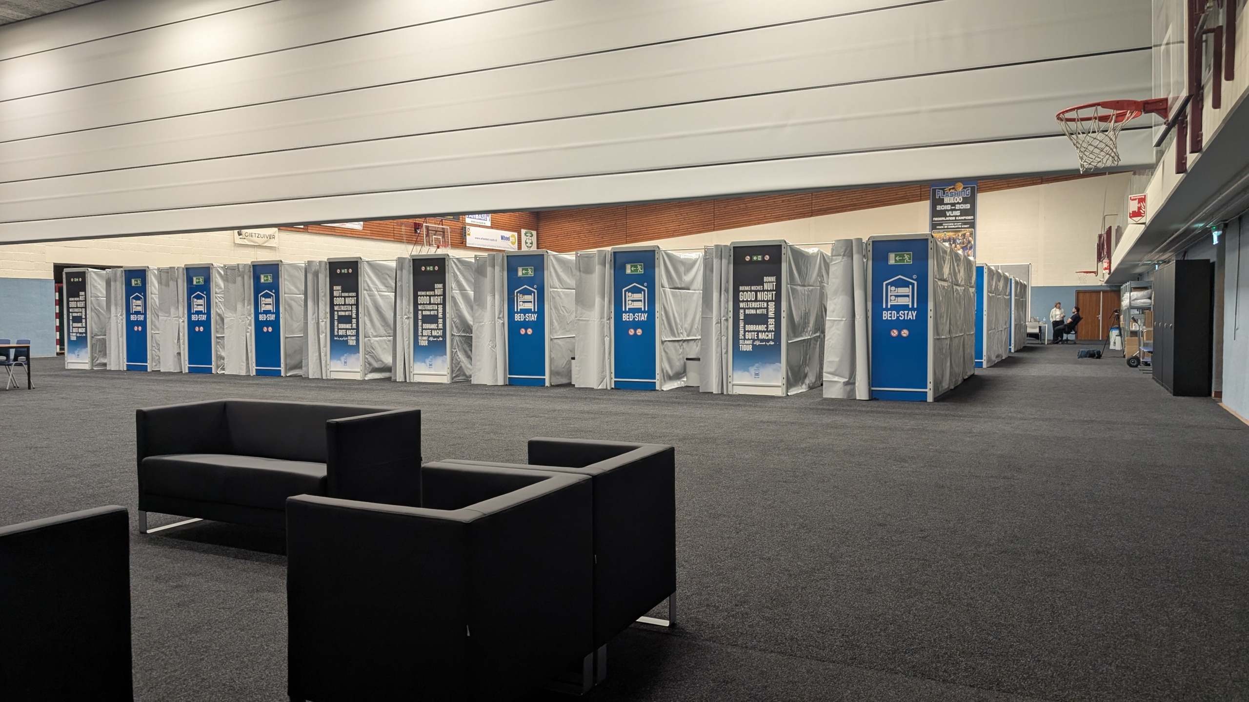 Rij met bed-cabines in een sporthal, enkele zwarte stoelen op de voorgrond, en een basketbalring aan de wand.