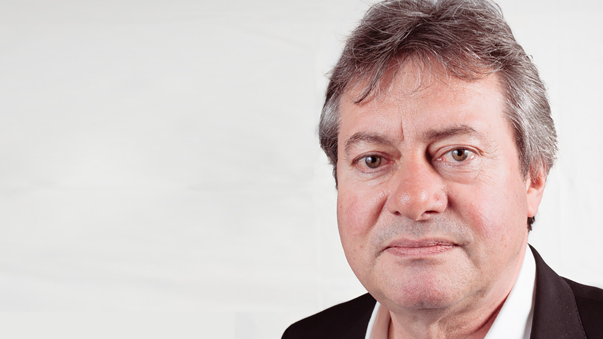 Close-up portret van een man met grijs haar in pak, tegen een lichte achtergrond