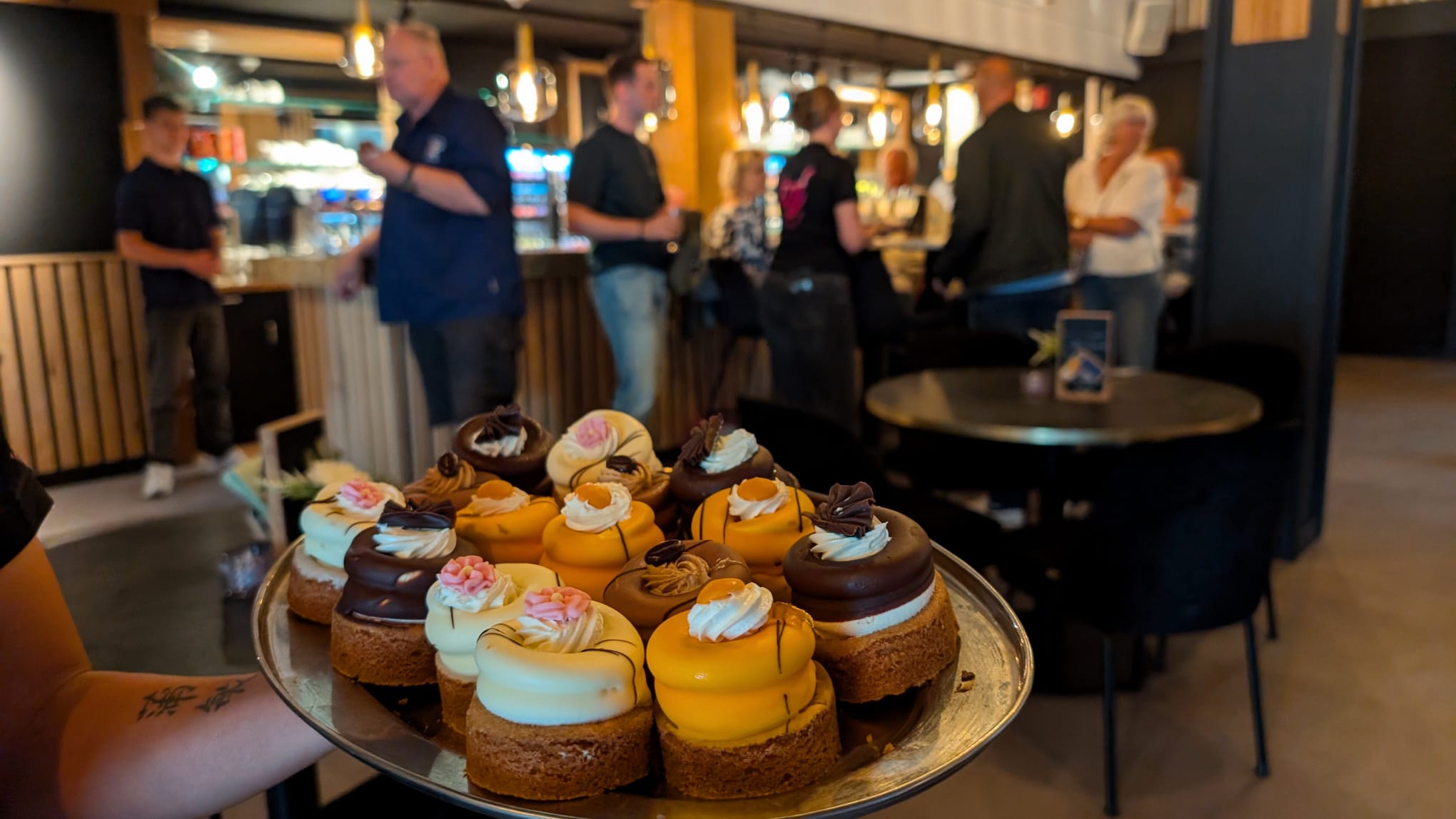 Een dienblad met verschillende kleurrijke gebakjes in een druk bezochte bar.