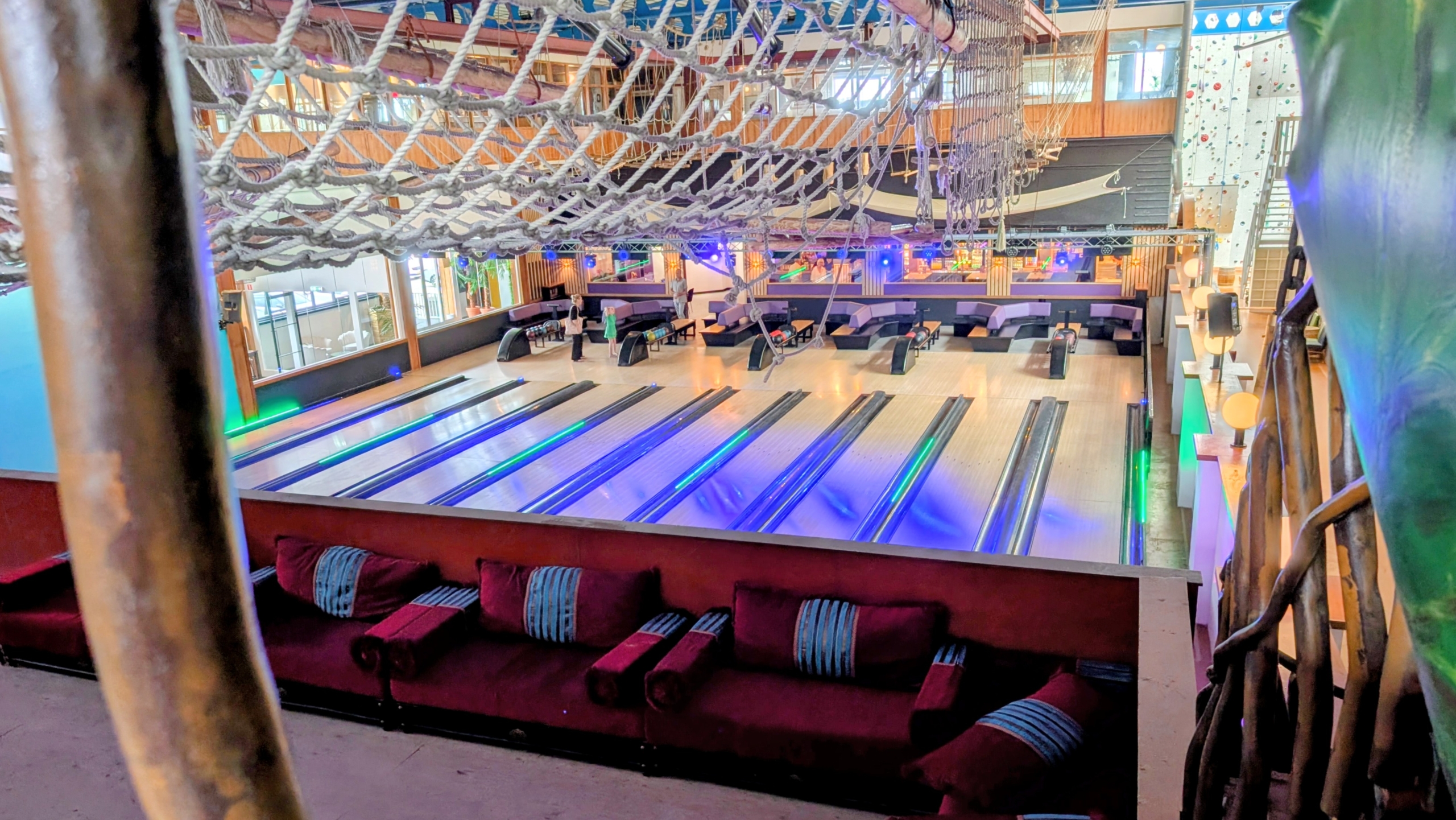 Een moderne bowlingbaan met kleurrijke verlichting en zitgedeelte, gezien vanaf een hoger gelegen plek met een klimnet erboven.