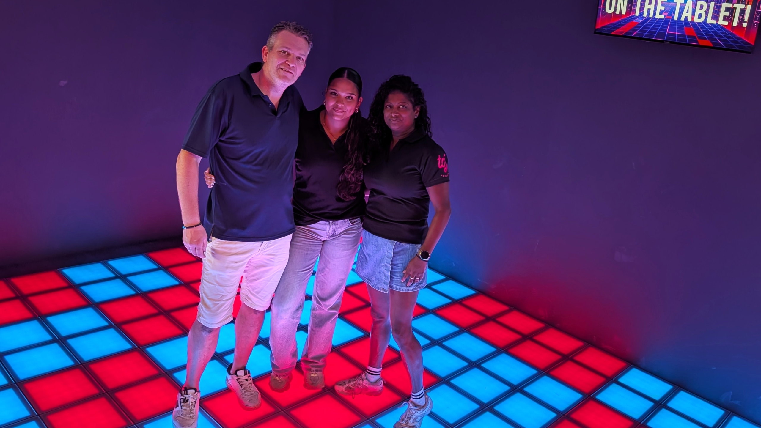 Drie mensen poseren samen op een vloer met rood en blauw verlichte tegels, tegen een paarse achtergrond.