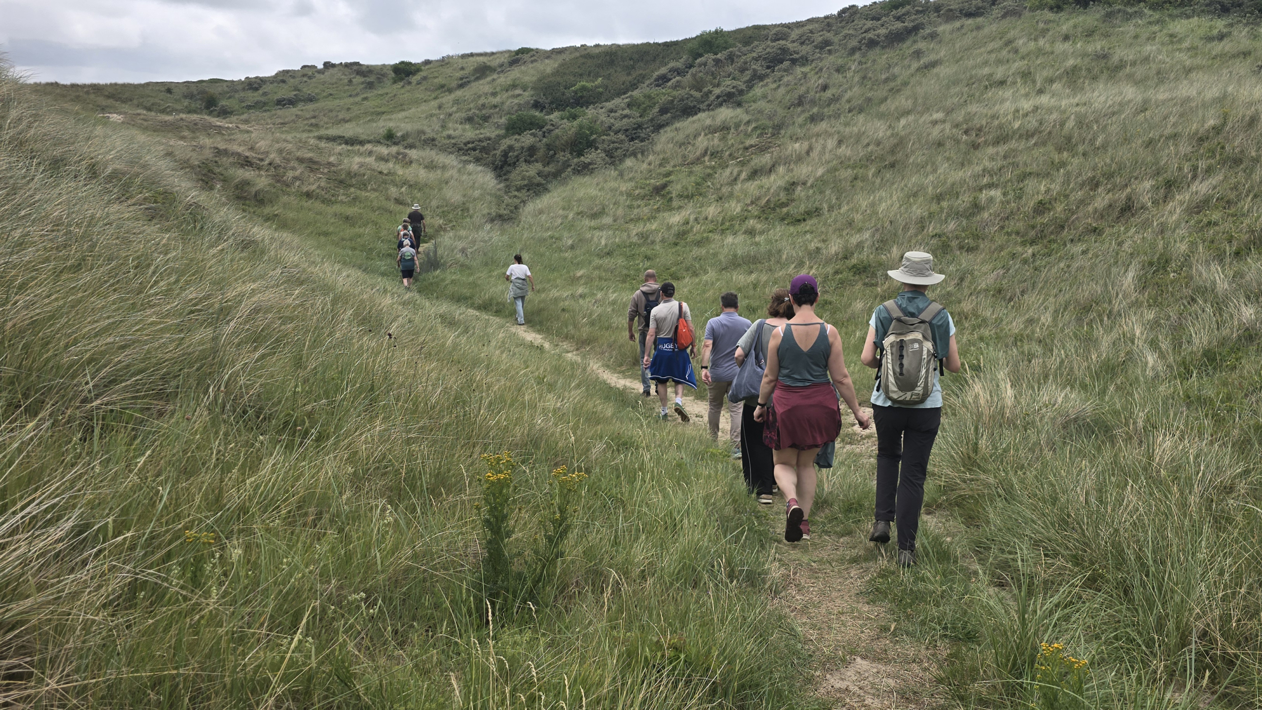 Een groep mensen wandelt over een smal pad door een heuvelachtig graslandschap onder een bewolkte lucht.