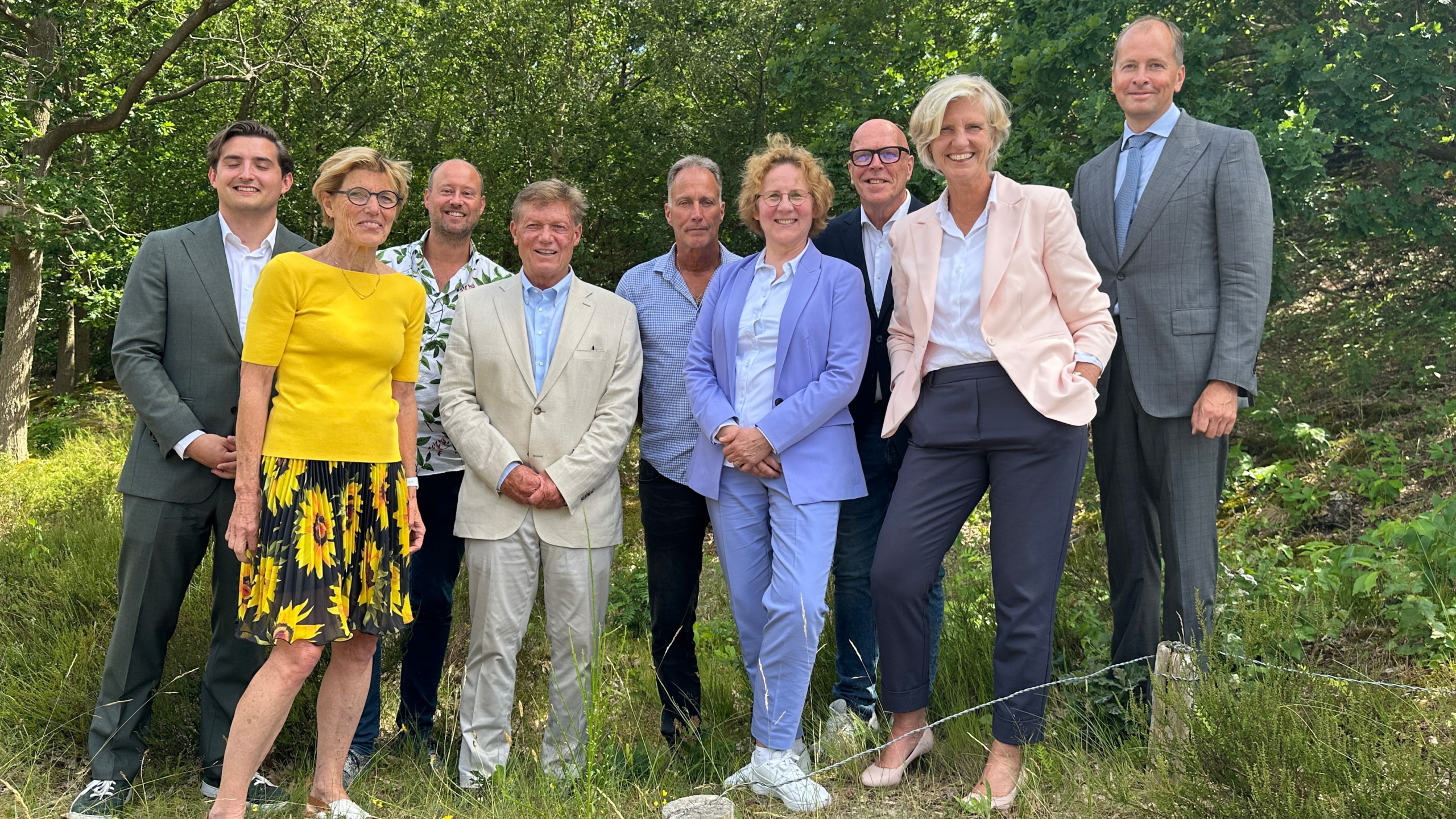 Een groep mensen poseert buiten, gekleed in formele kleding en lachend voor de camera omringd door bomen.