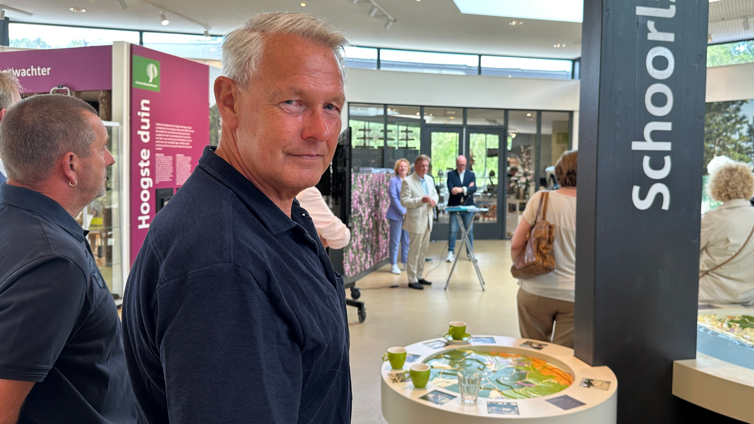 Een man staat in een tentoonstelling met een paneel waarop "Schoorl" en "Hoogste duin" staat; mensen zijn in gesprek op de achtergrond.
