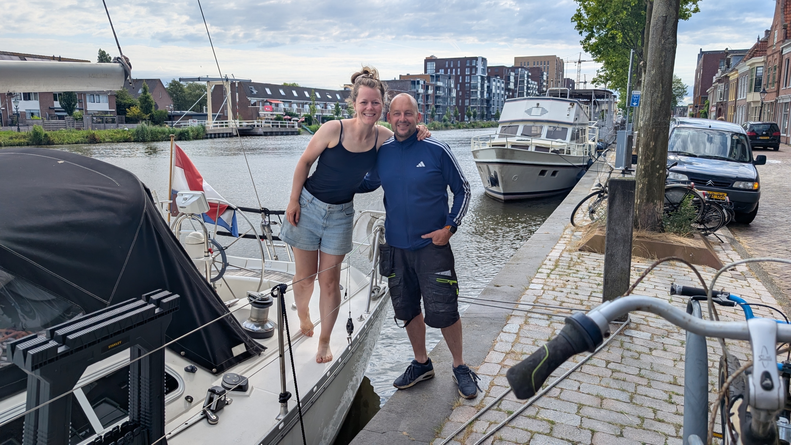 Twee mensen staan glimlachend op een zeilboot aan een kade, met boten en gebouwen op de achtergrond.